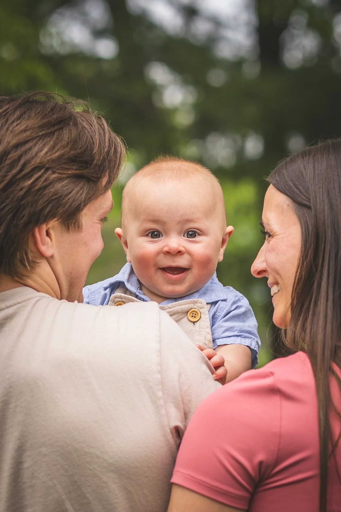 A baby being held by a man and a woman, with the baby smiling and looking directly at the camera. The man is on the left, the woman on the right, both facing each other. The background is blurred greenery.