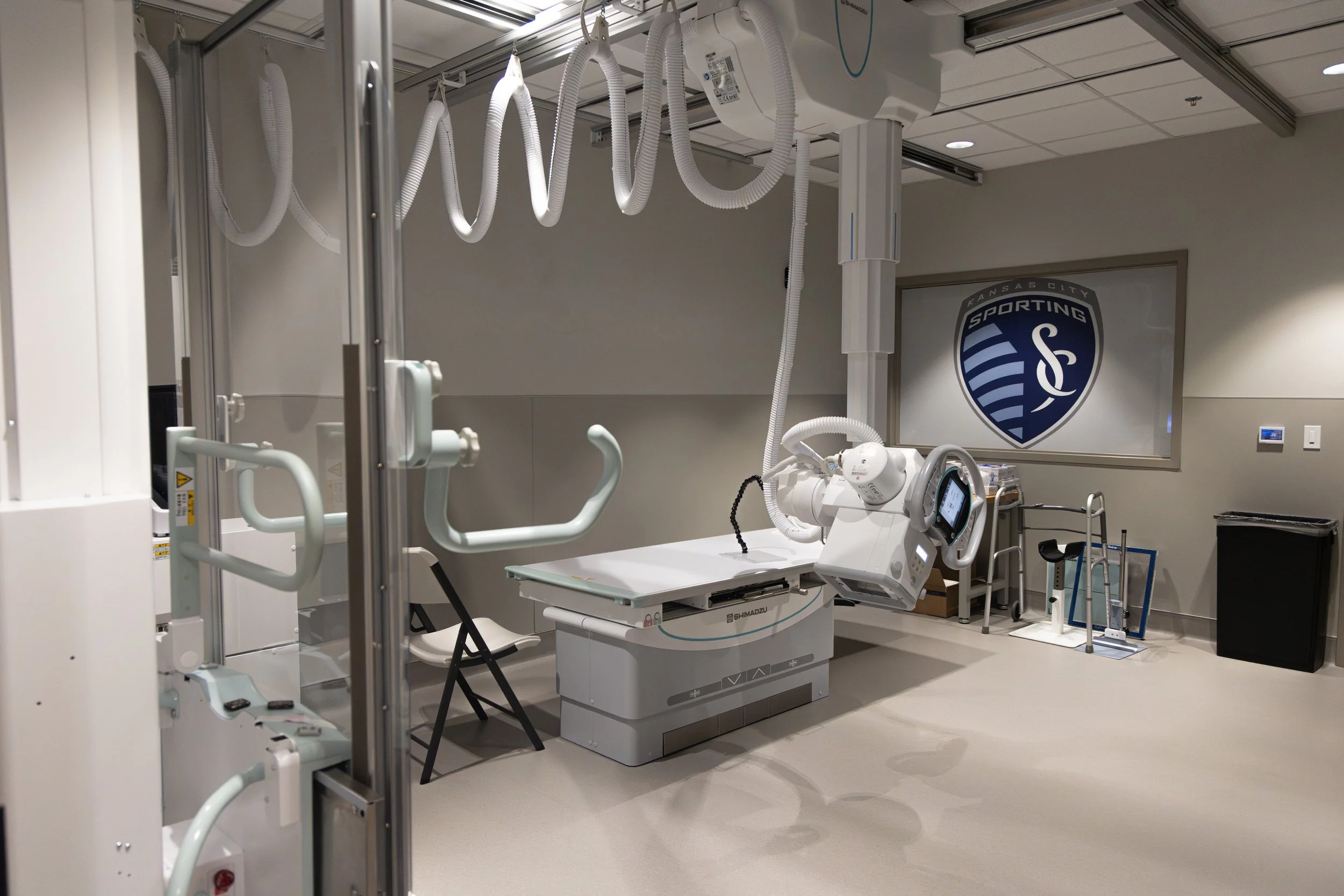 Hospital room with medical equipment, including a robotic surgical device, monitors, and IV hooks, with a Kansas City Sporting logo on the wall.