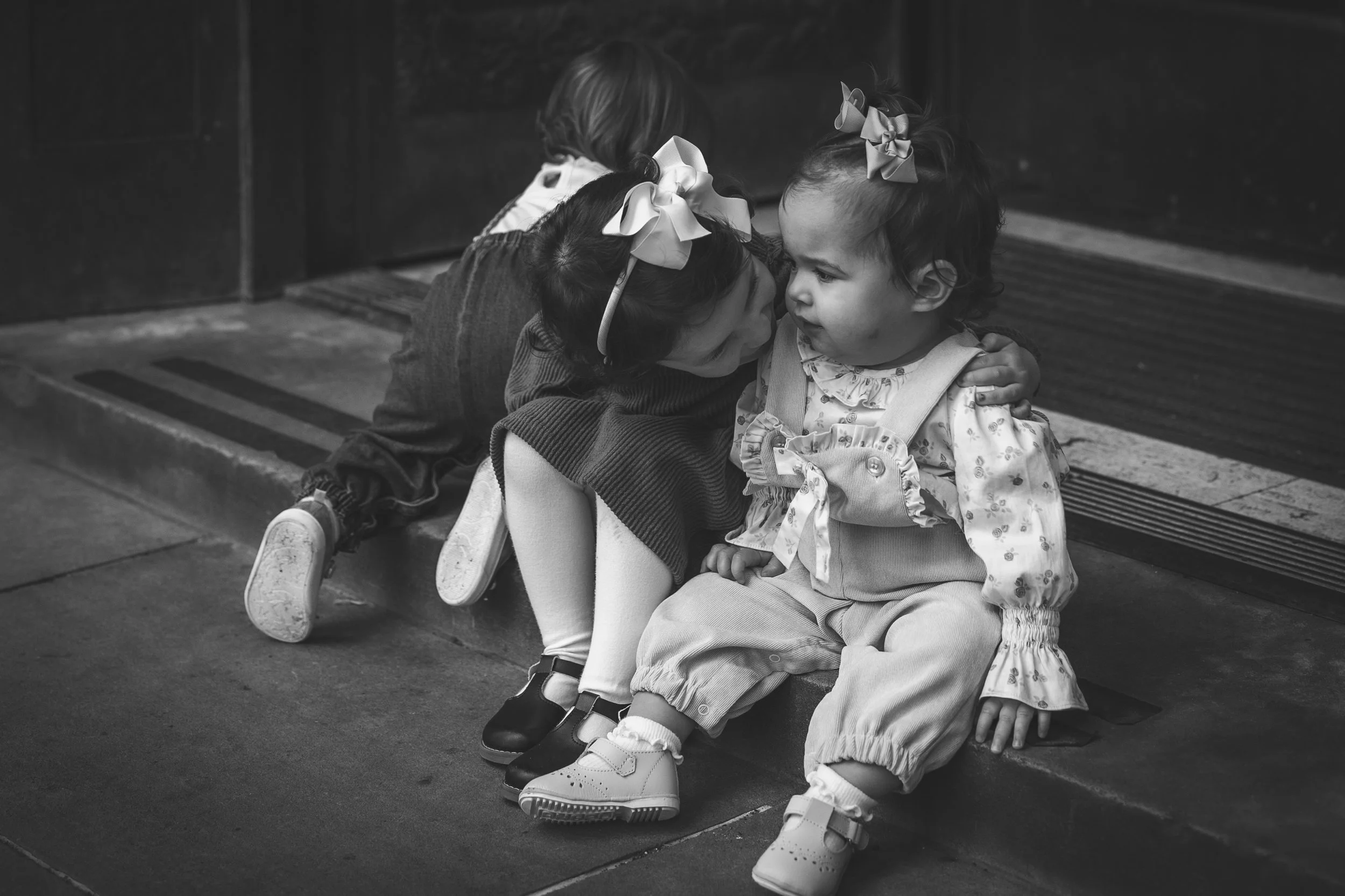 Two young girls sitting on a sidewalk, with the older girl leaning in to kiss the younger girl on the cheek, both wearing bows in their hair.