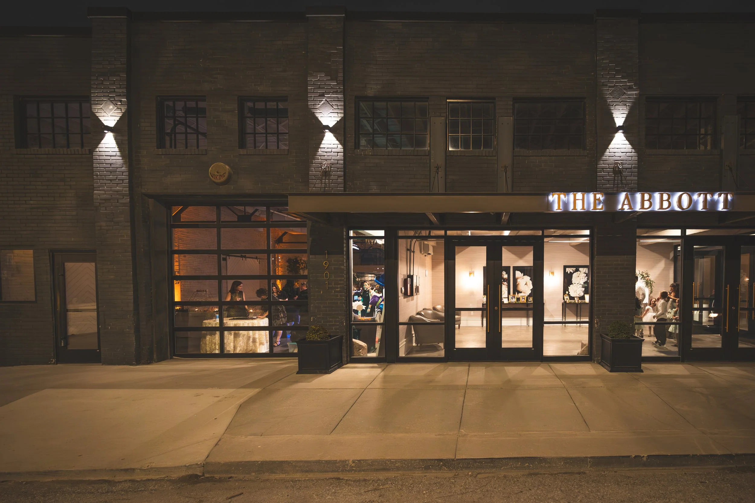 Night view of restaurant entrance called 'The Abbott' with illuminated sign and large glass doors, interior with people dining visible through windows, brick exterior, decorative lighting, and sidewalk in front.