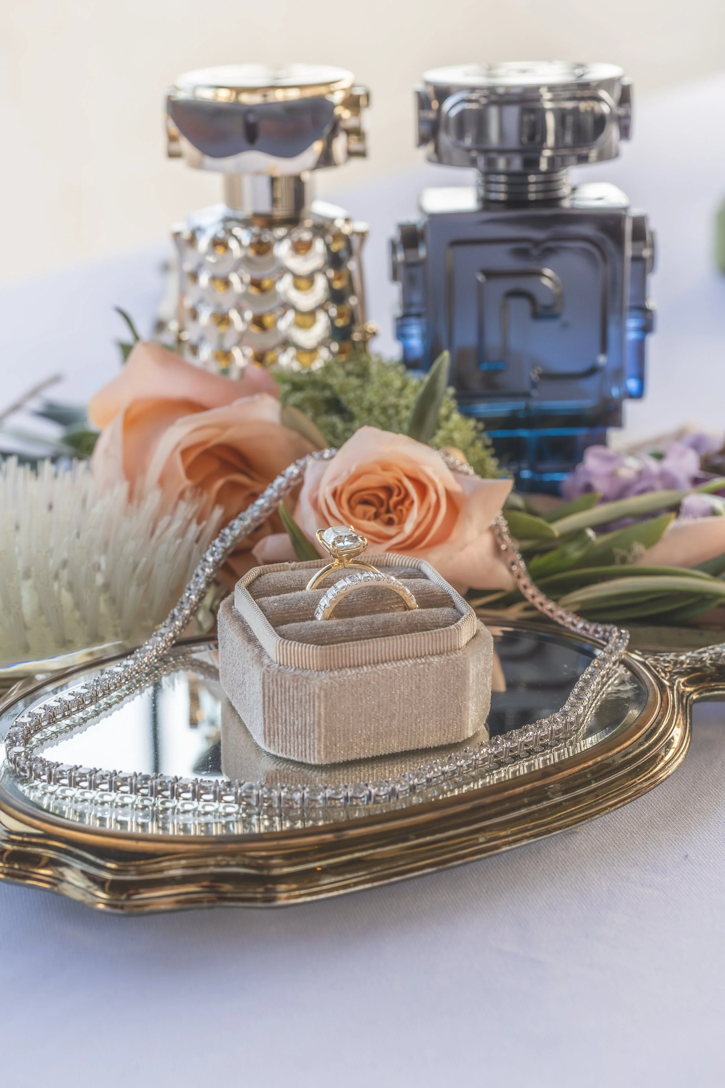 A silver tray holds jewelry, including a sparkling diamond ring in a beige velvet ring box, a silver chain, and a pair of earrings, with a floral arrangement featuring peach roses and other flowers in the background.