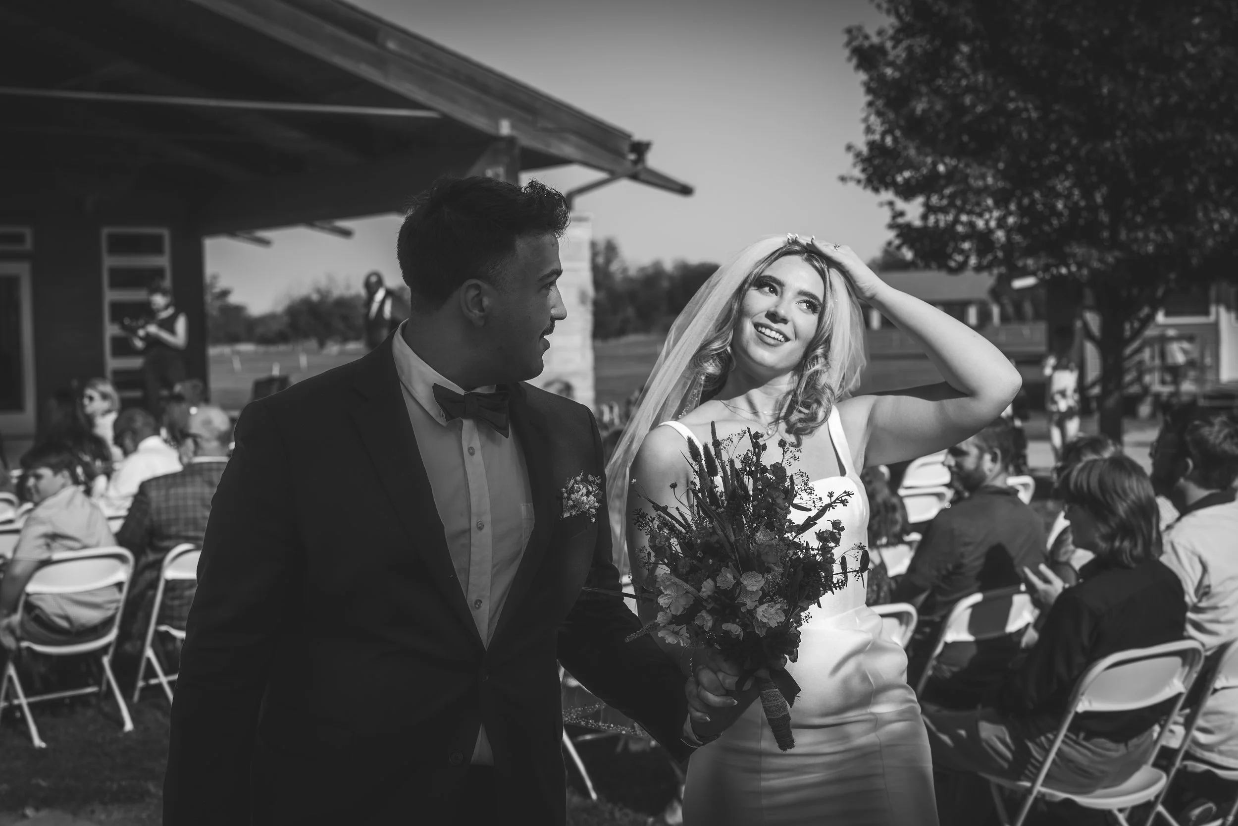 A black and white photo of a bride and groom holding hands, walking outdoors during a wedding ceremony. The bride is smiling and adjusting her hair with her left hand, wearing a wedding dress and veil, holding a bouquet. The groom is dressed in a tux