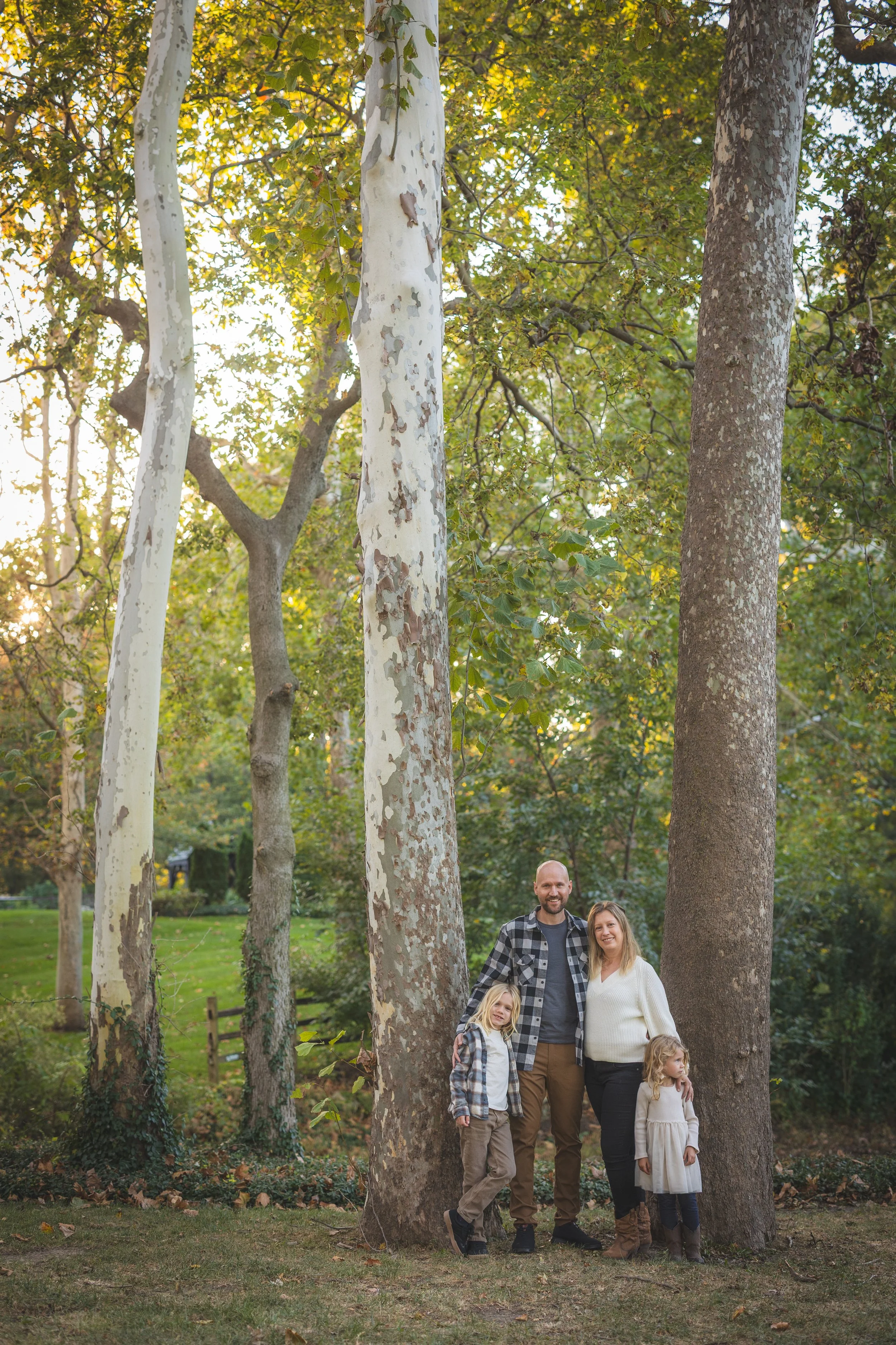 A family of five standing outdoors among tall trees in a park during daytime. The family includes two adults and three children, all smiling and dressed casually. The background features green grass, trees, and a fence.