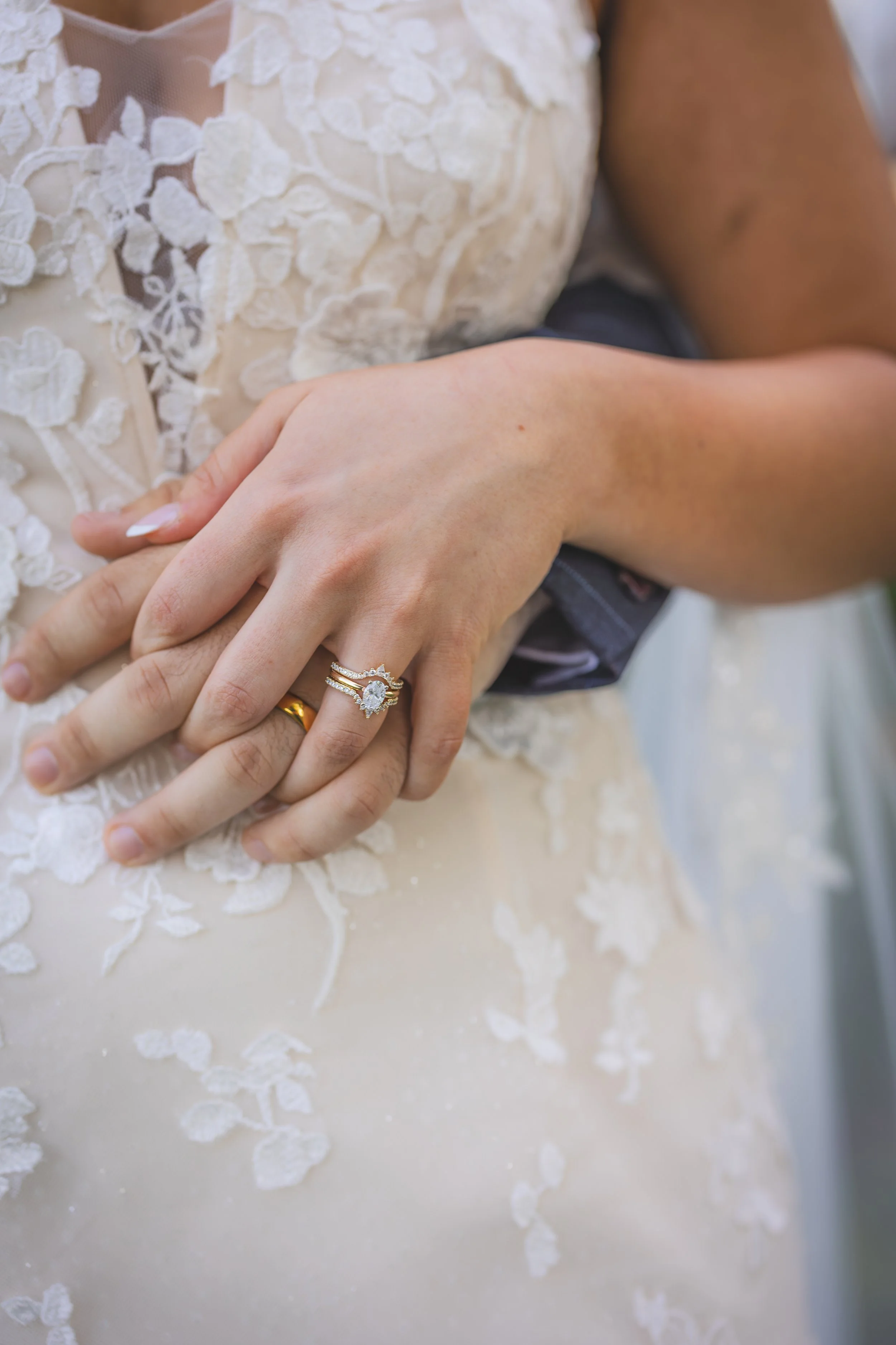 Close-up of two hands with wedding rings, one hand resting on top of the other, showing a wedding band and an engagement ring, in front of a dress with lace details.