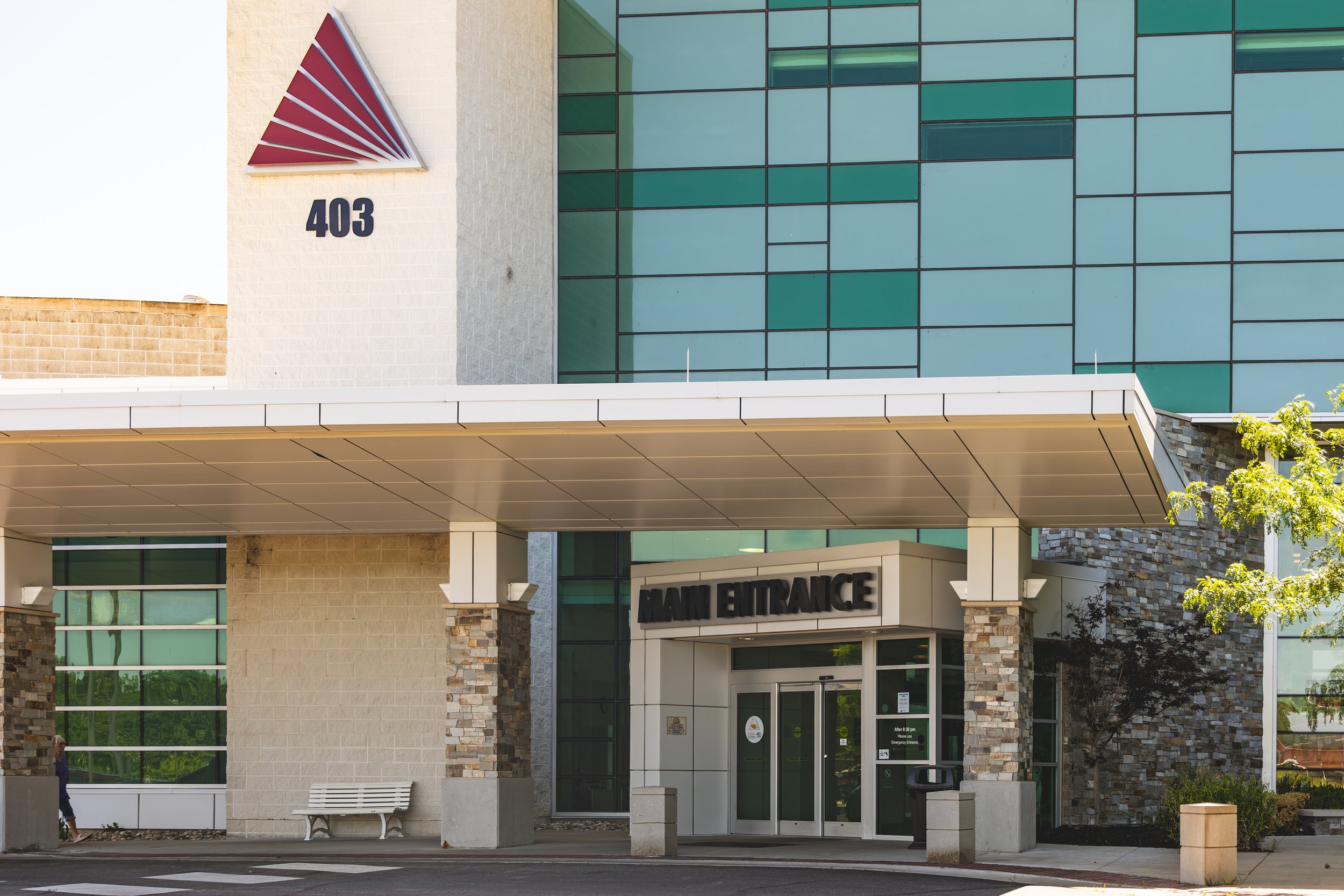Exterior of a building with a sign that reads 'Main Entrance,' featuring large glass windows, brick and stone architectural elements, and the number 403 on the wall with a red and white logo above it.