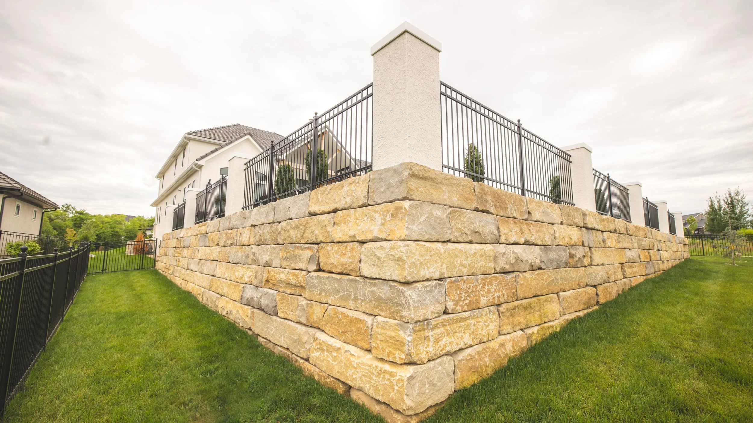 A raised stone patio with a metal railing surrounding it in the backyard of a suburban home, with the house and trees in the background.