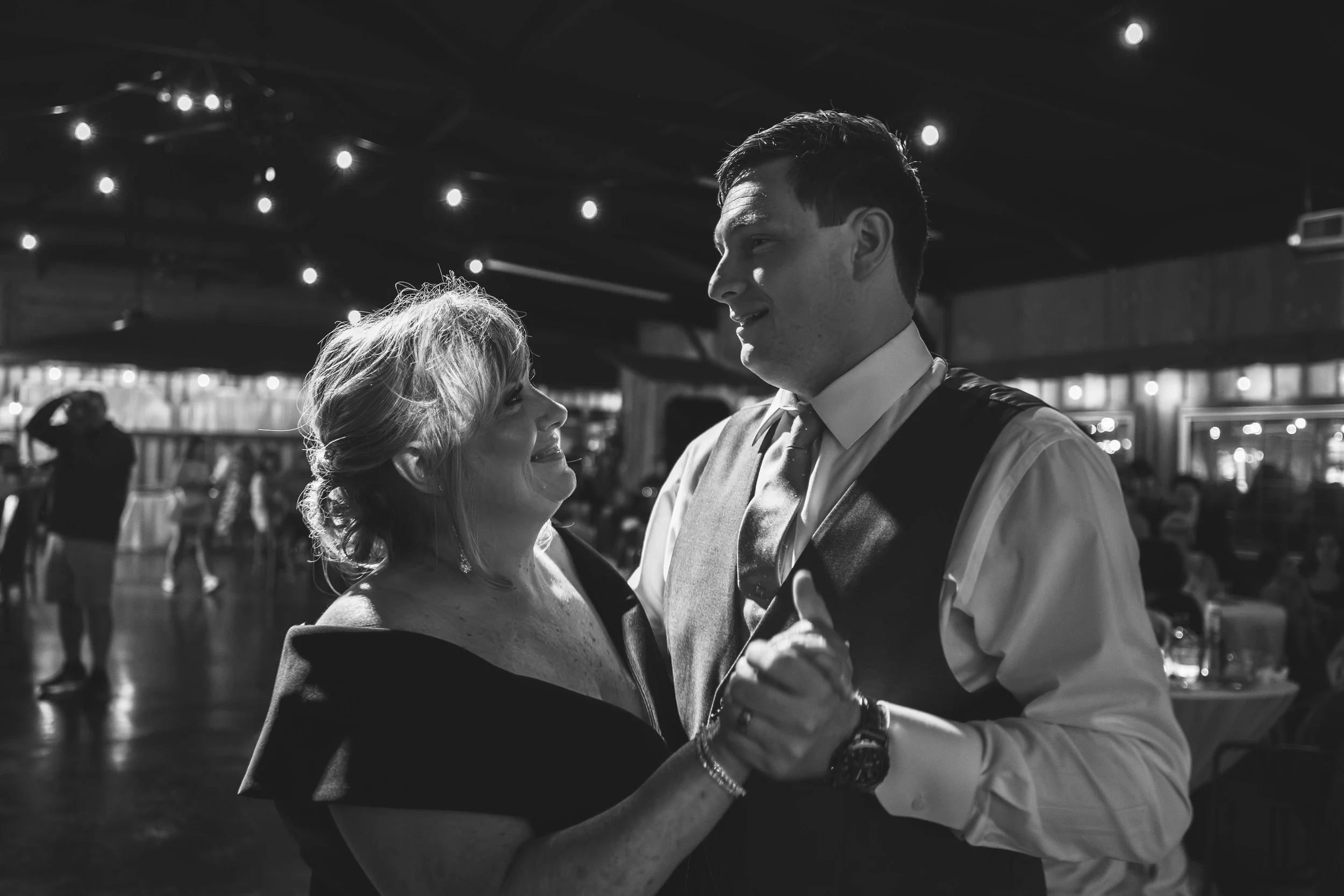 A man and woman are dancing and smiling at each other in a dimly lit venue with string lights overhead.