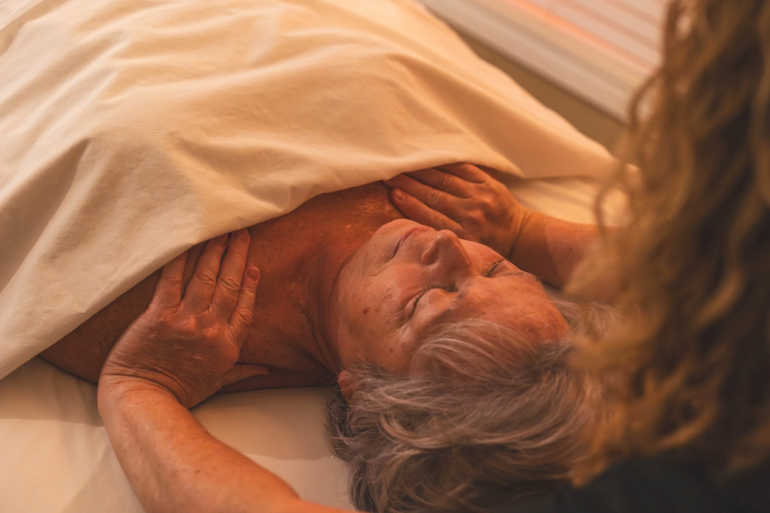 An elderly woman lying on a bed with her eyes closed, covering her chest with a white sheet, with a person's hand gently touching her shoulder.