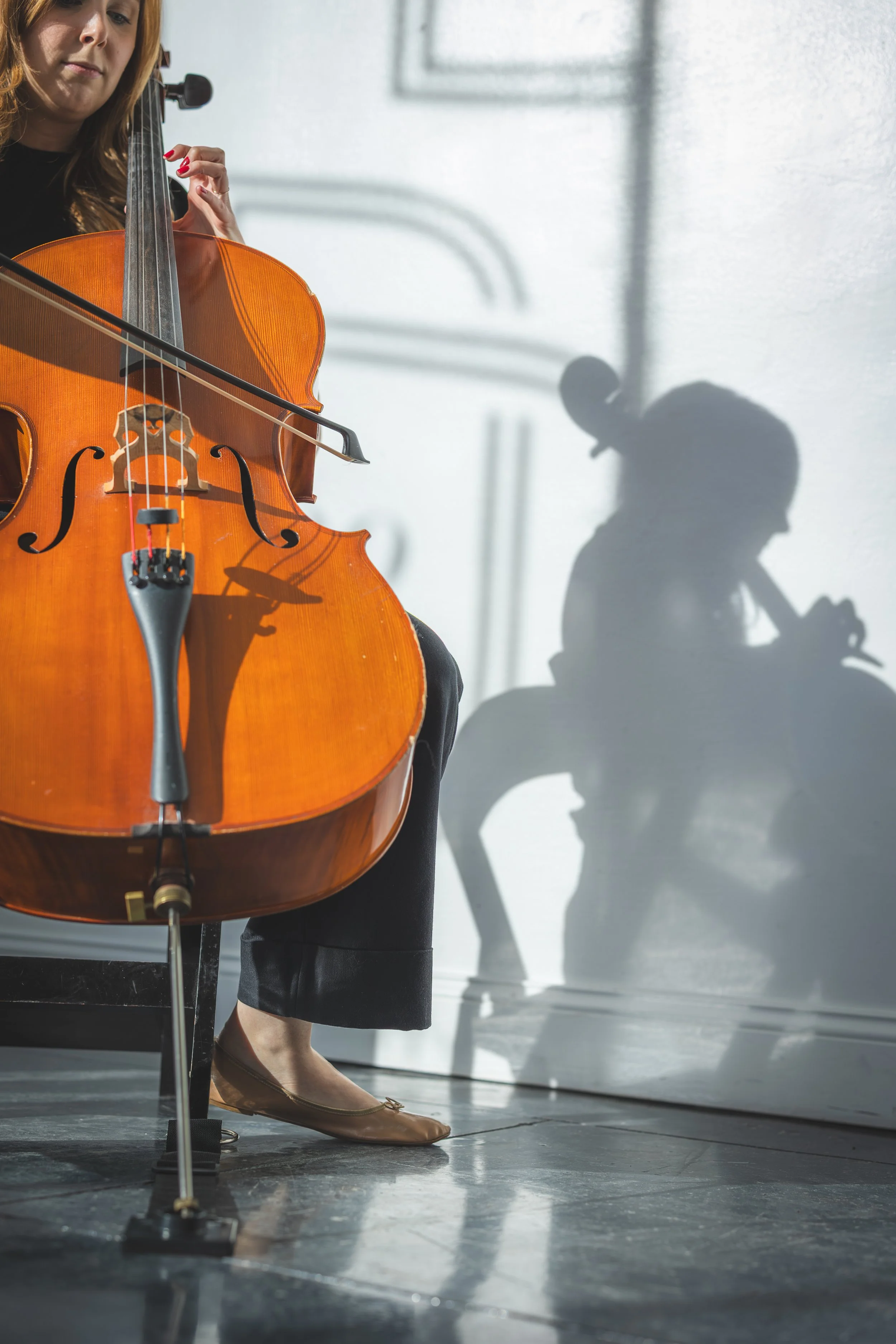 A woman playing a cello in a room with white walls and a large shadow of herself on the wall. She is dressed in a black outfit and brown shoes, and her face is partially visible. The focus mostly on the cello and the shadow.