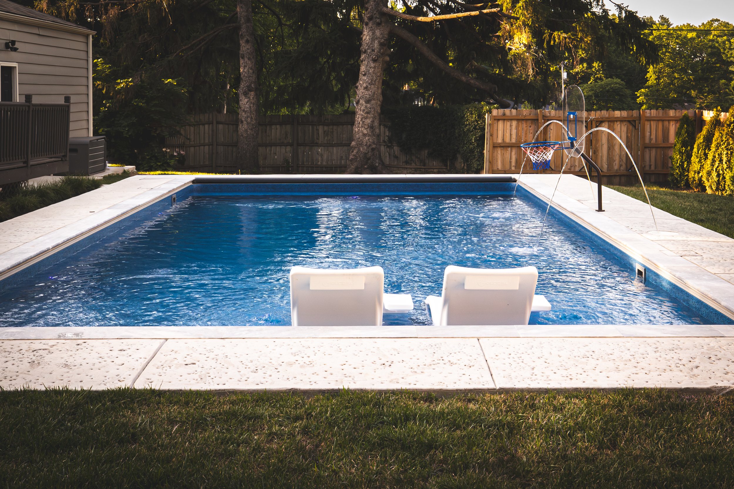 A backyard swimming pool with two white lounge chairs in the foreground, a basketball hoop over the pool, surrounded by a concrete deck and wooden fencing, with trees and greenery in the background.