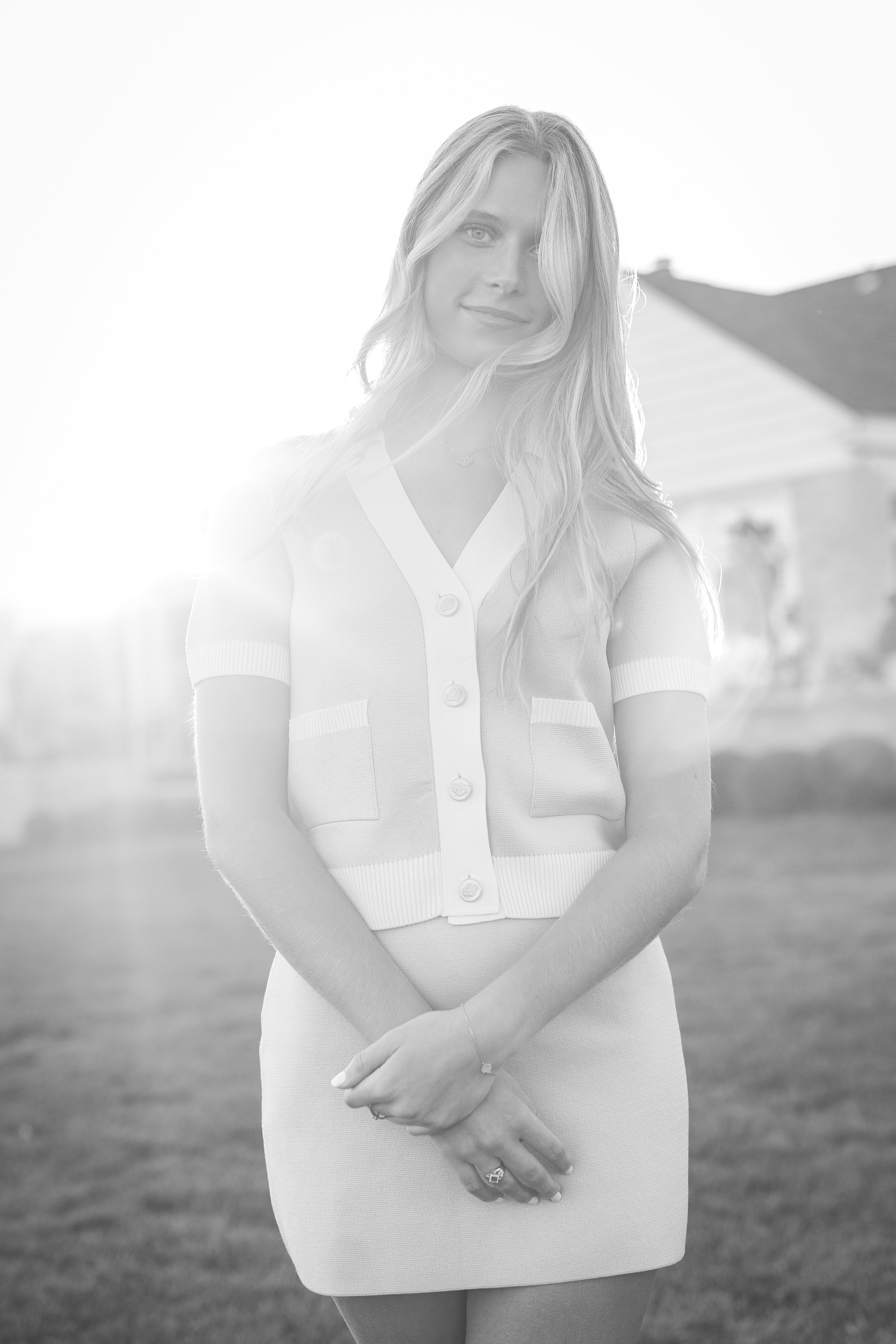 Black and white photo of a woman standing outdoors with a house in the background, smiling, with her hair styled in loose waves, wearing a short-sleeved button-up cardigan and a skirt.