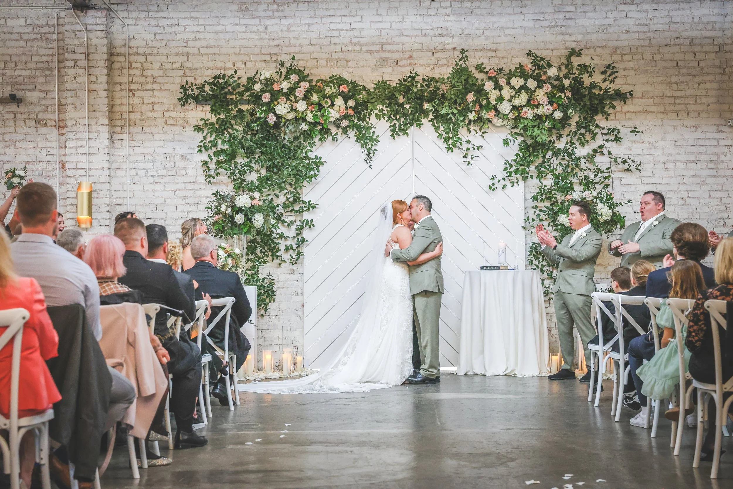 A wedding ceremony with a bride and groom kissing, surrounded by guests, in front of a floral backdrop and white wall, with officiants clapping and celebrating.