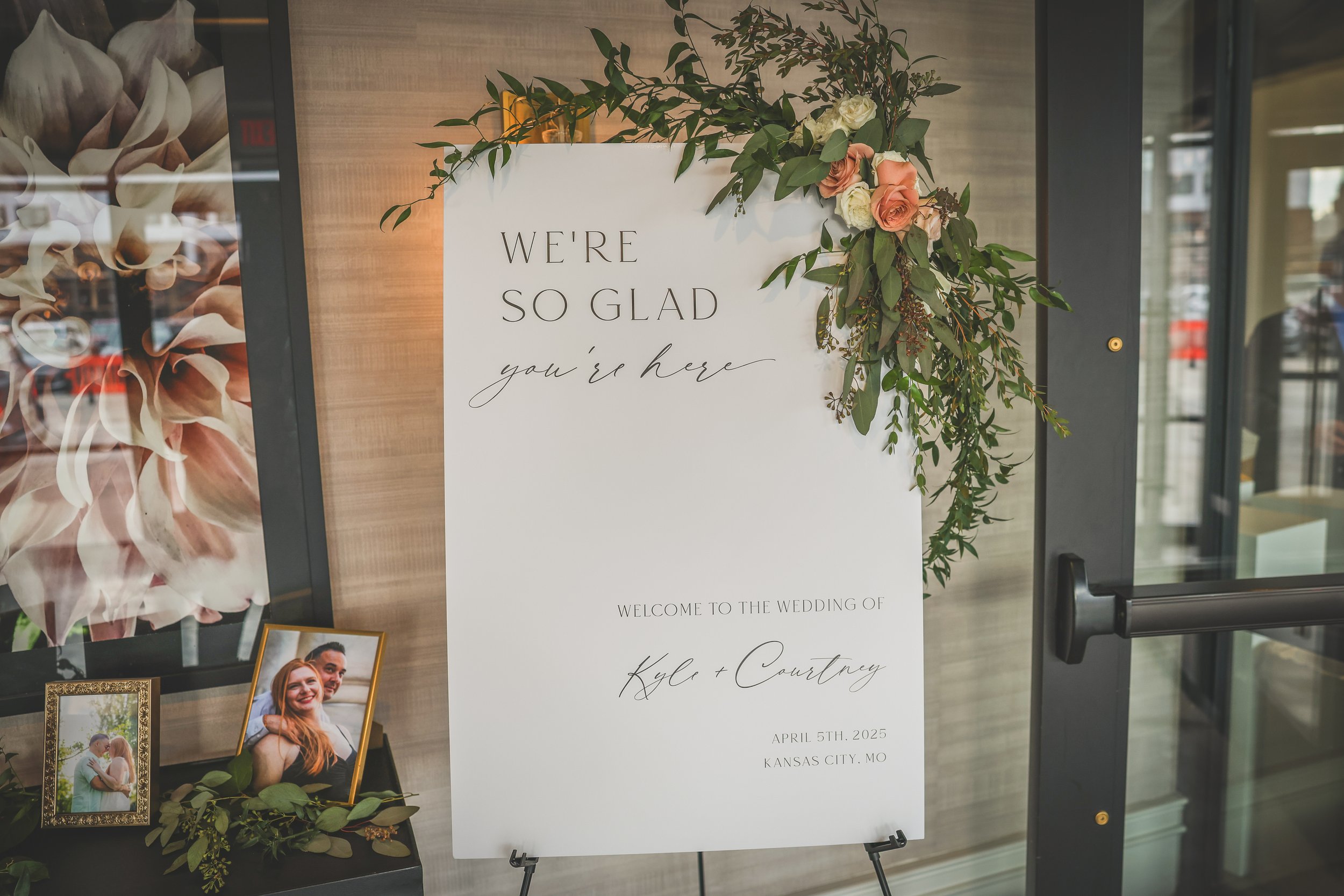 A wedding sign with floral decorations reads, 'We're so glad you're here,' welcoming guests to Kyle and Courtney's wedding on April 5th, 2025, in Kansas City, MO.