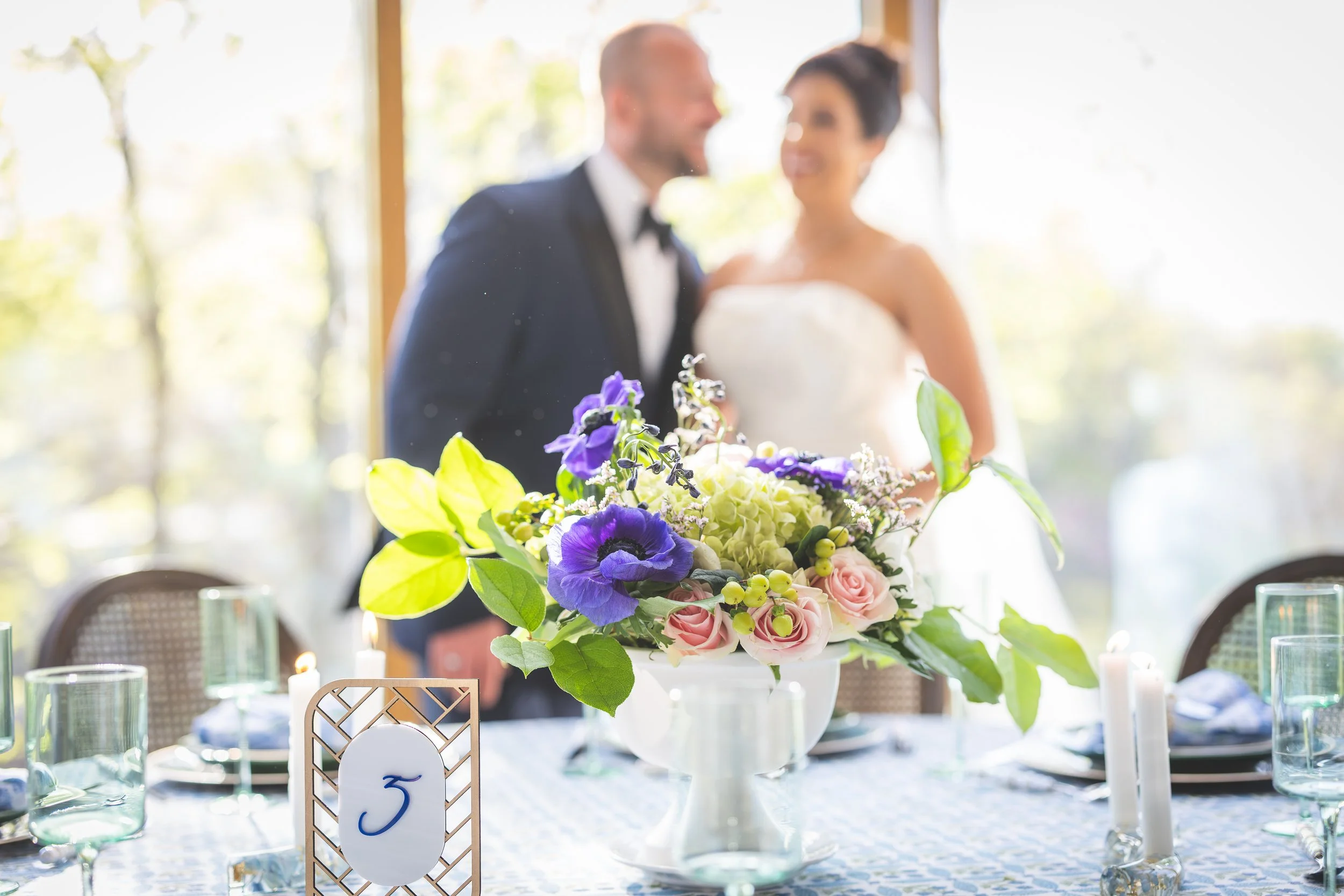 A wedding reception table with a floral centerpiece in a white vase, candles, and tableware in front of a blurred bride and groom standing and talking in the background.