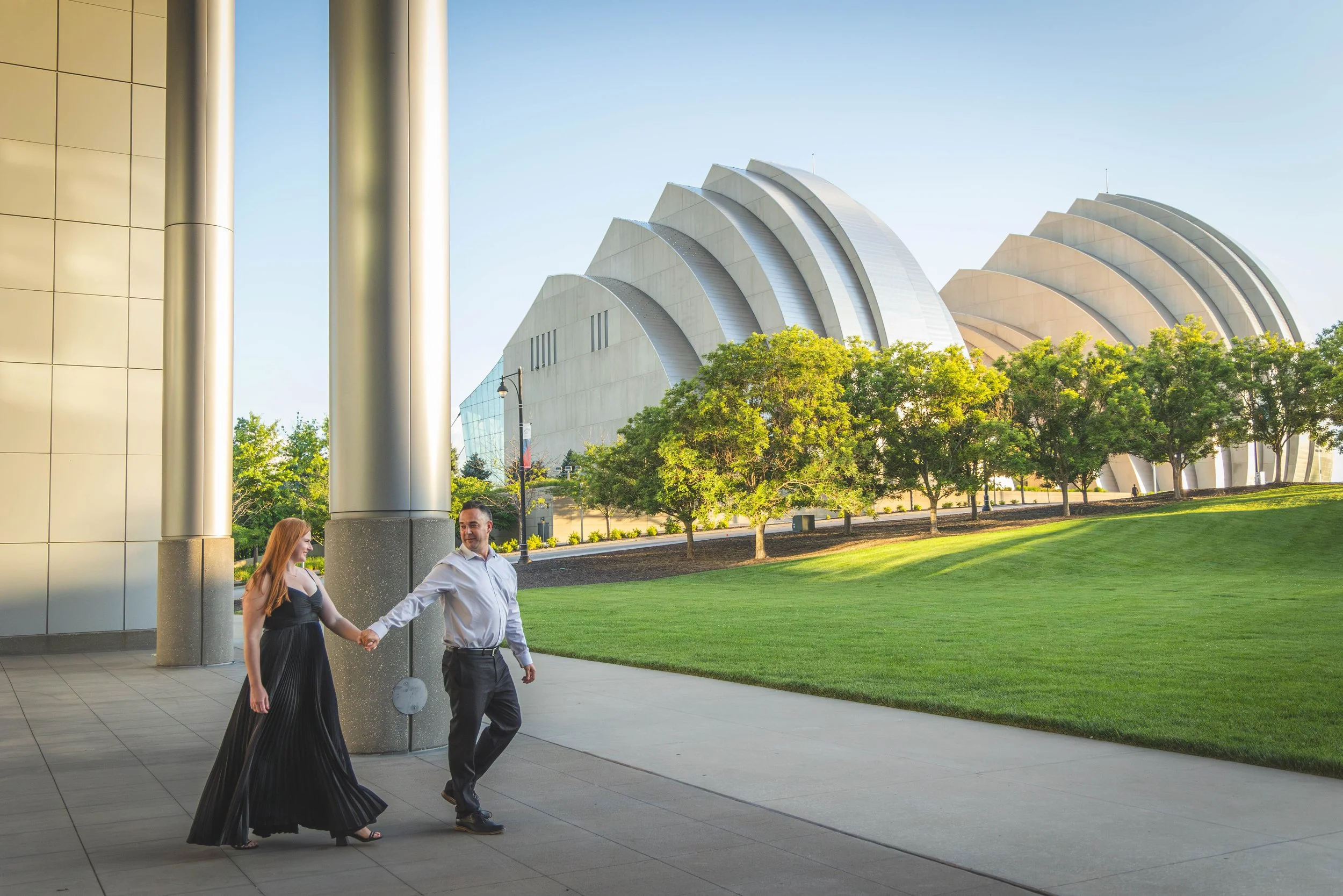 A couple holding hands and walking outside a modern building with curved white roofs and lush green trees.