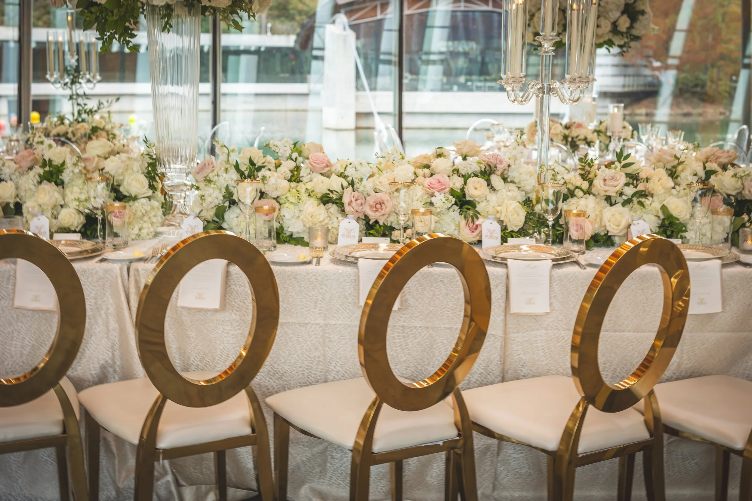 Elegant banquet table decorated with white and pale pink roses and greenery, set with gold-rimmed plates, glassware, and gold candles, with gold circular chair backs in the foreground and large glass vases and chandeliers overhead, in a bright room w