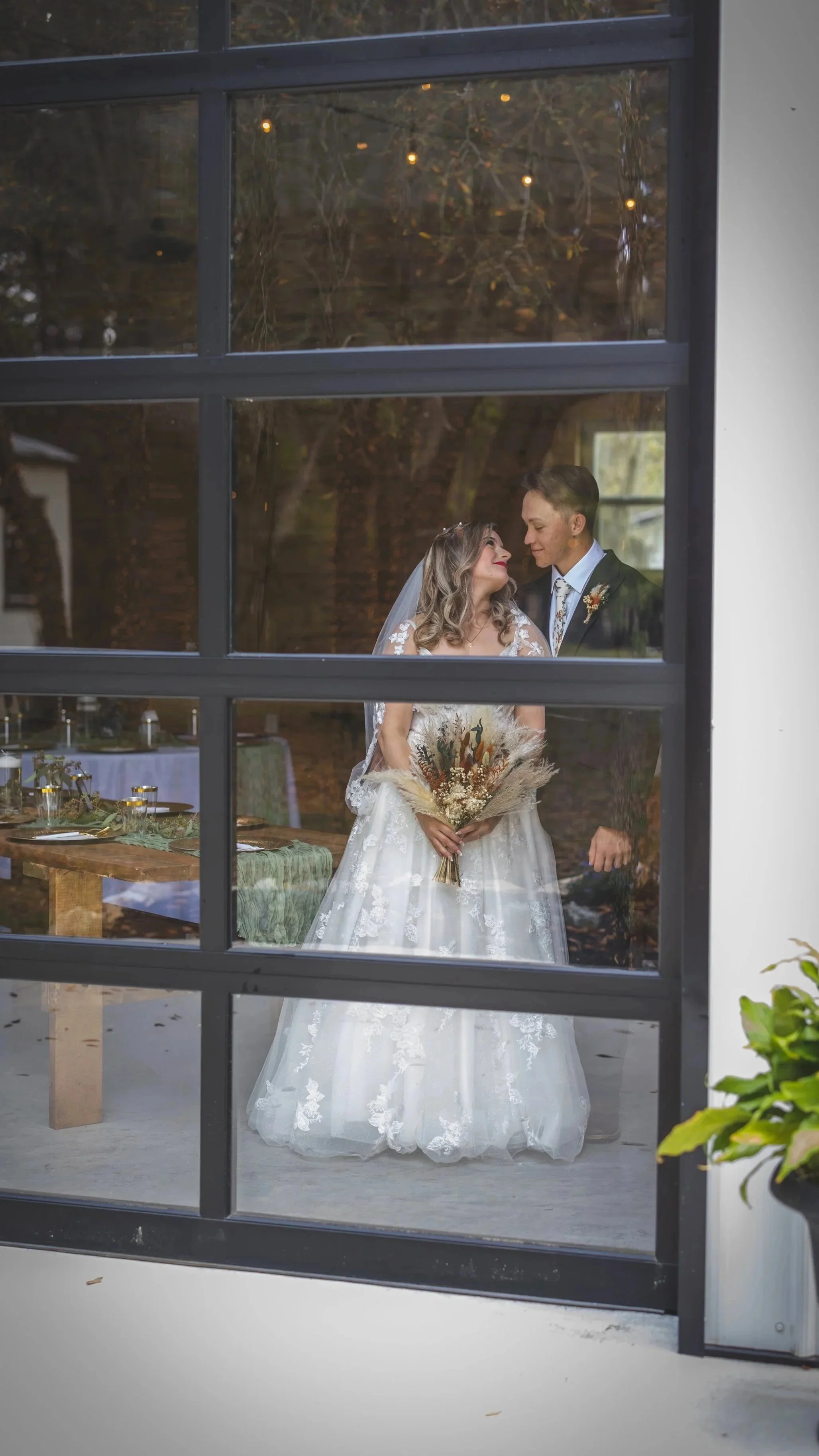 A bride and groom looking at each other through a large window during their wedding celebration. The bride is holding a bouquet and wearing a white lace wedding gown, while the groom is dressed in a suit with a boutonniere. The interior behind them i