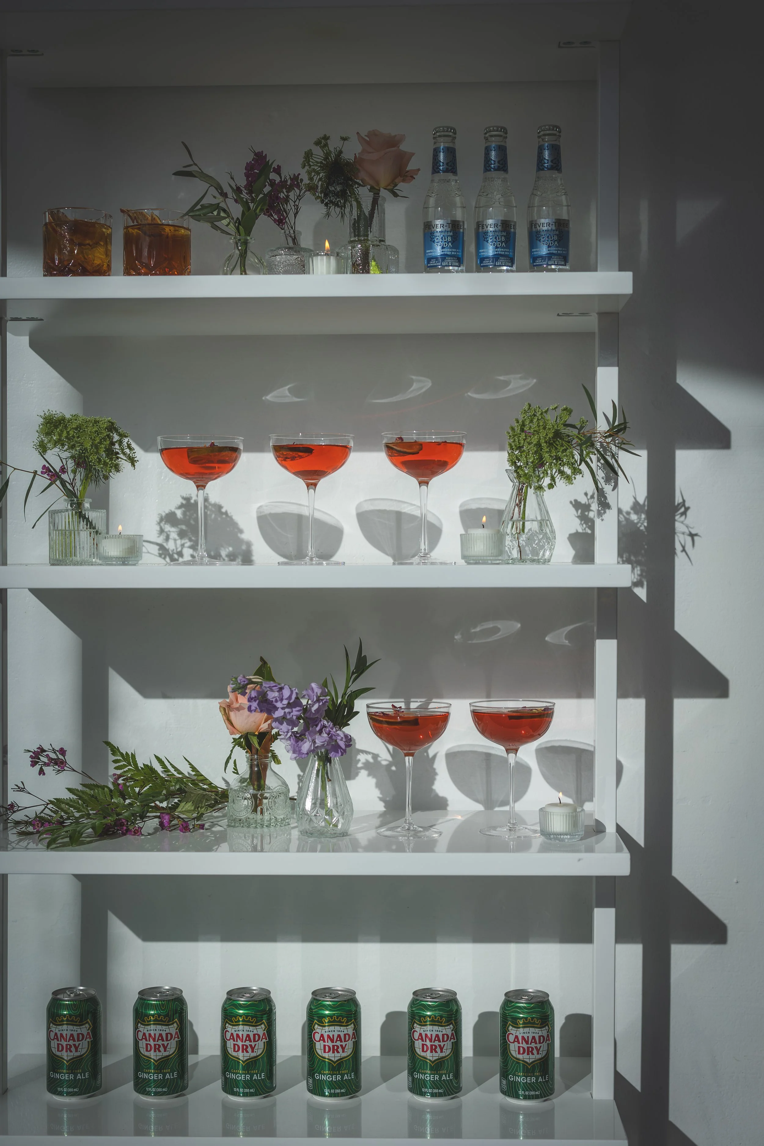 A white shelf displaying various drinks, flowers, and candles. The top row has two glasses of amber-colored liquid, a small bouquet, and three bottles of Fever-Tree tonic water. The middle row has three glasses of red-colored beverage, and bottles of