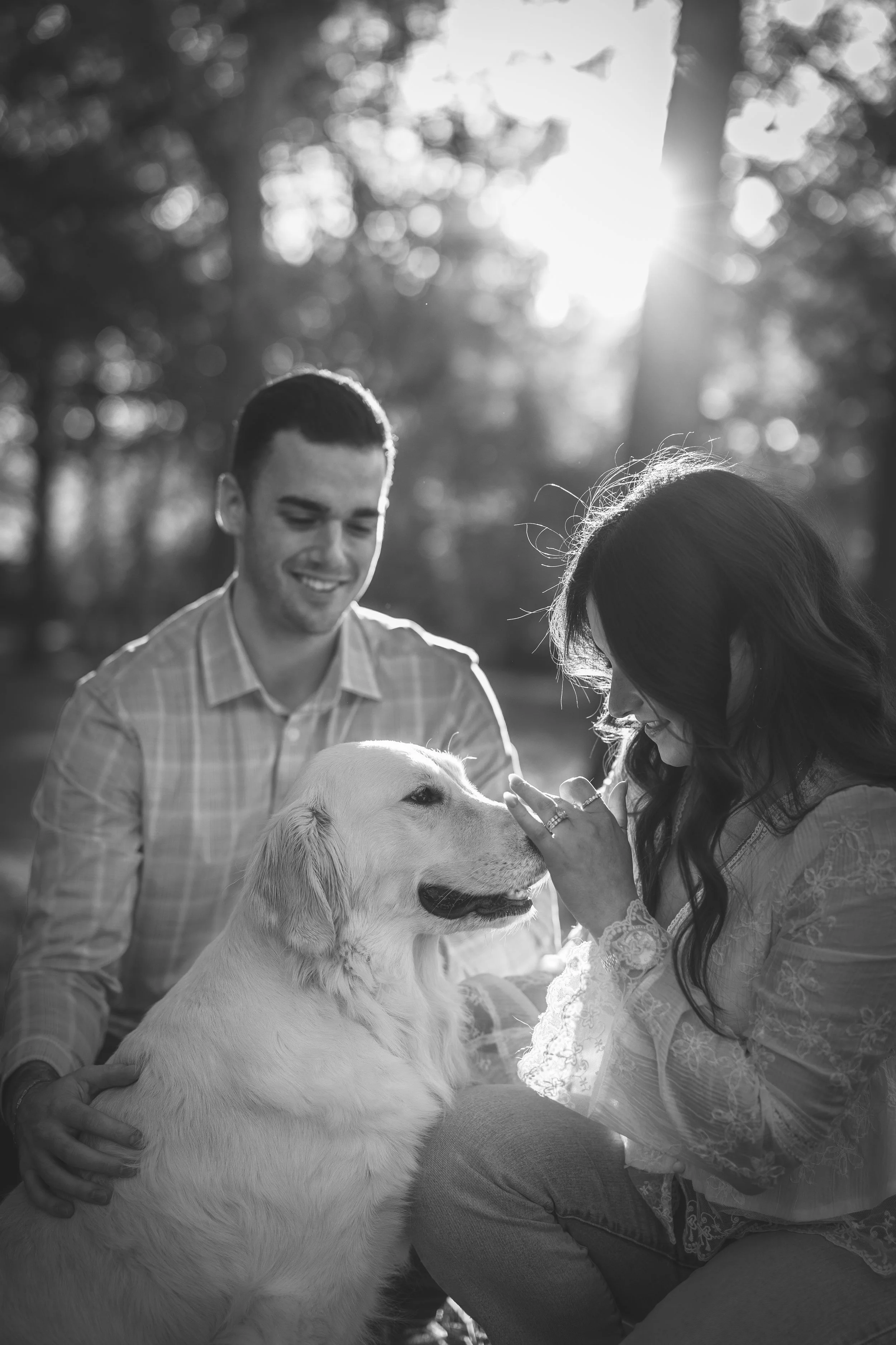 A black and white photo of a woman, a man, and a golden retriever dog outdoors. The woman is petting the dog, which is sitting and smiling. The man is standing behind the woman, smiling, with trees and sunlight in the background.