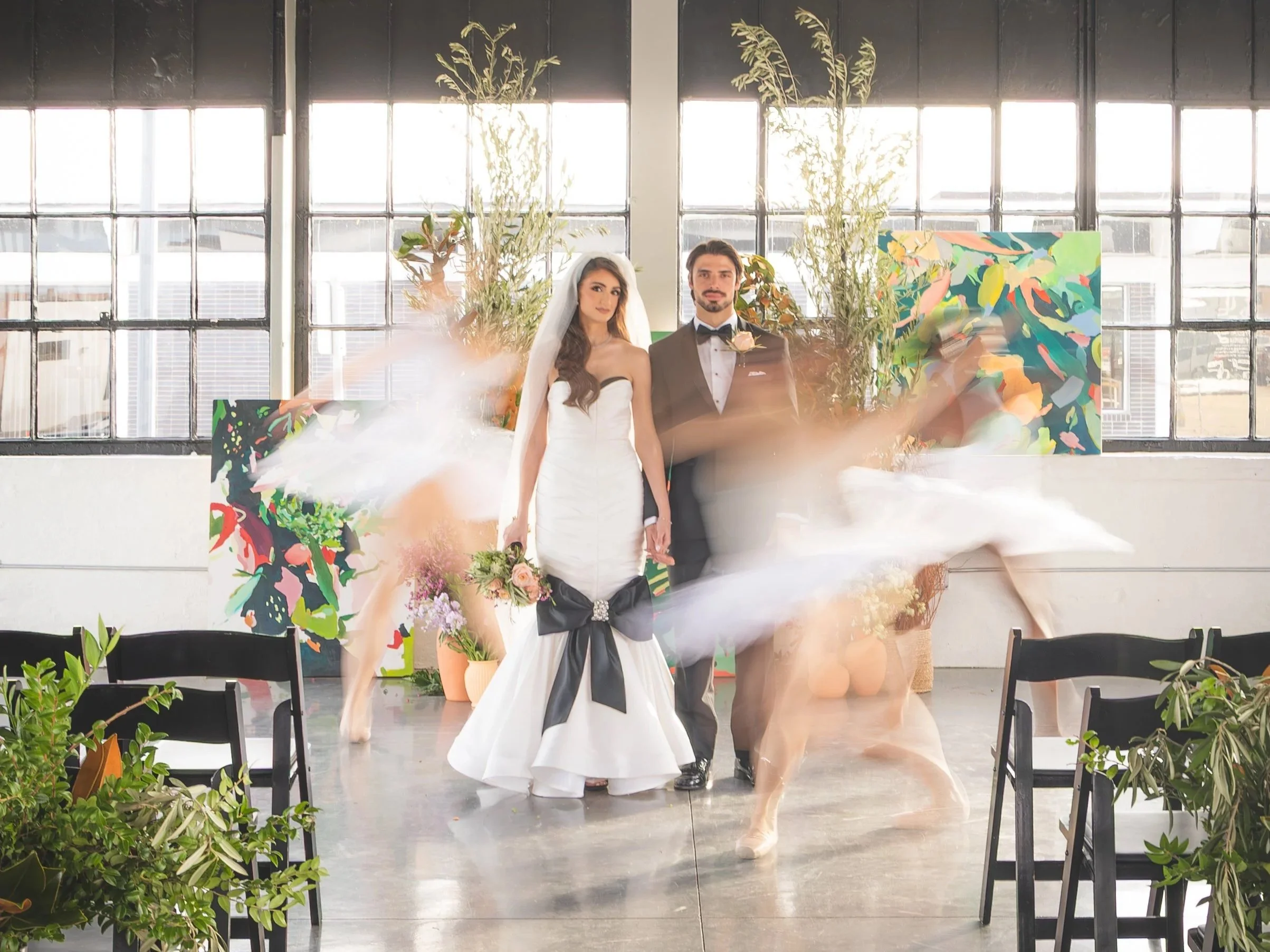 A bride and groom standing together in a brightly lit industrial-style space with large windows, surrounded by dancers in motion, colorful abstract art, and various plants and flowers.