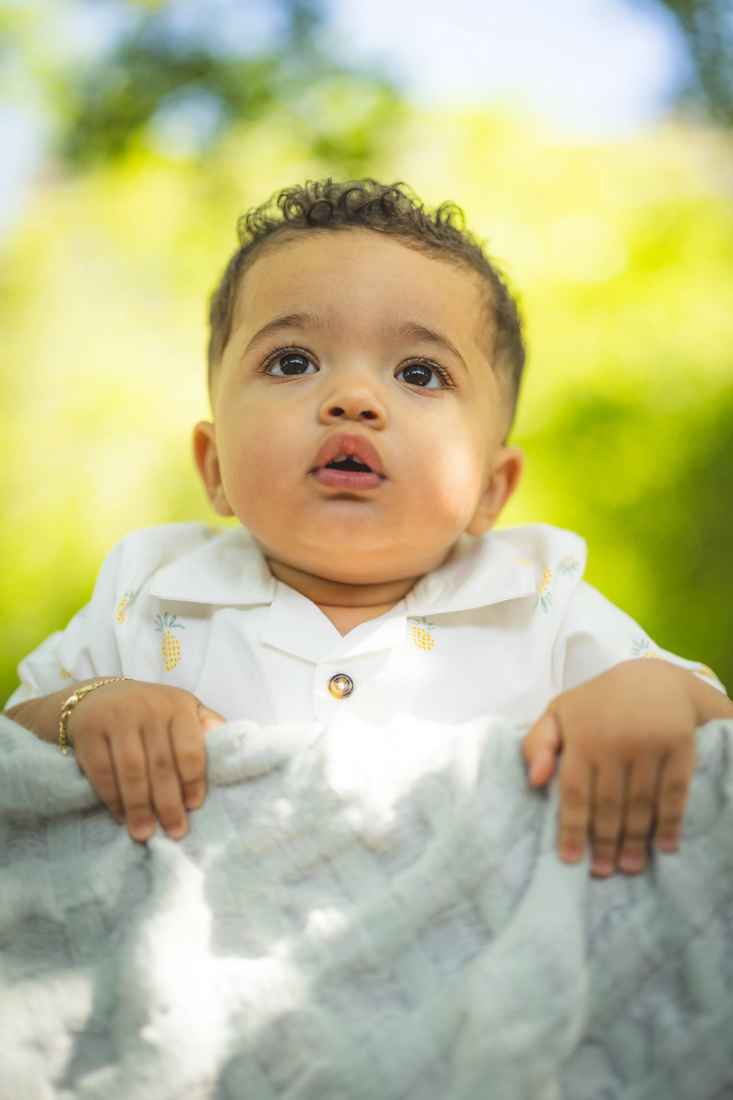 Close-up of a young child with curly hair holding onto a blanket outdoors against a blurred green and blue background.