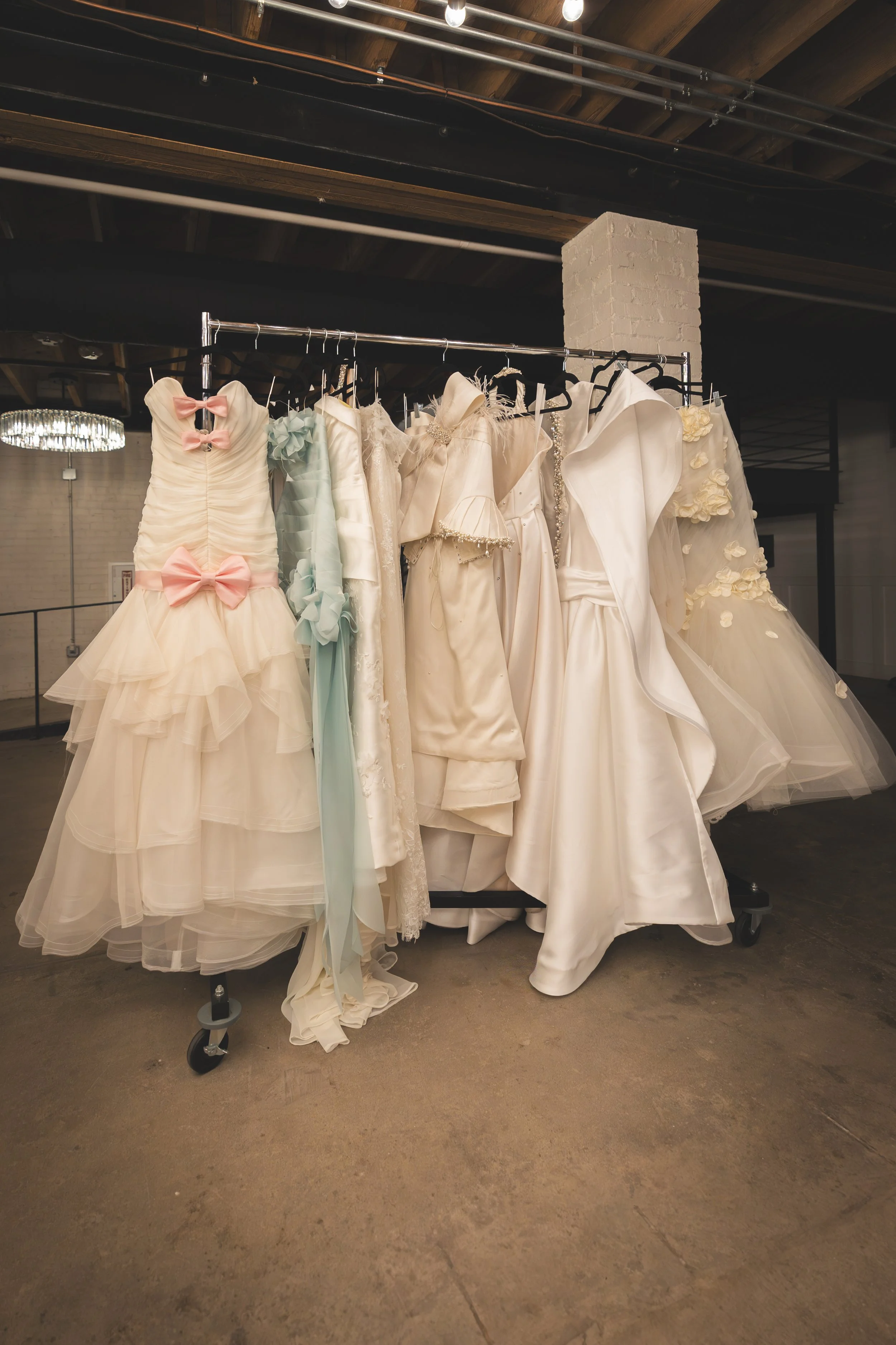 Clothing rack with wedding dresses hanging in an industrial-style room with exposed wooden beams and brick pillar.