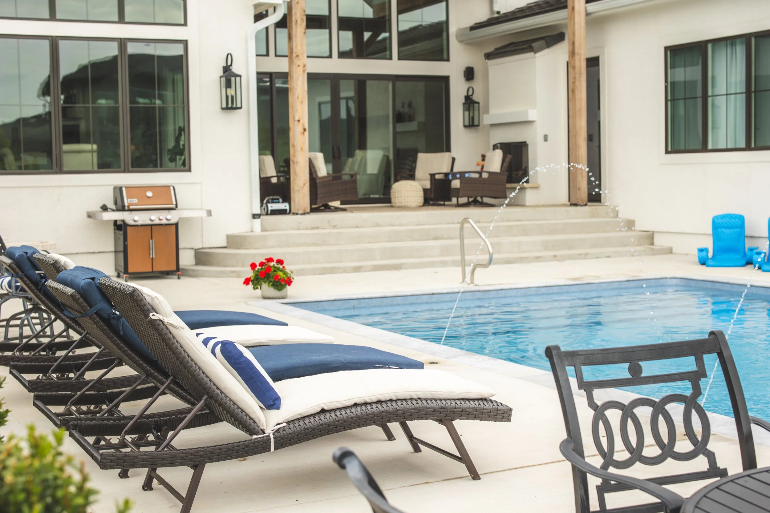 View of a backyard pool area with lounge chairs, outdoor furniture, and a house with large windows and a patio.