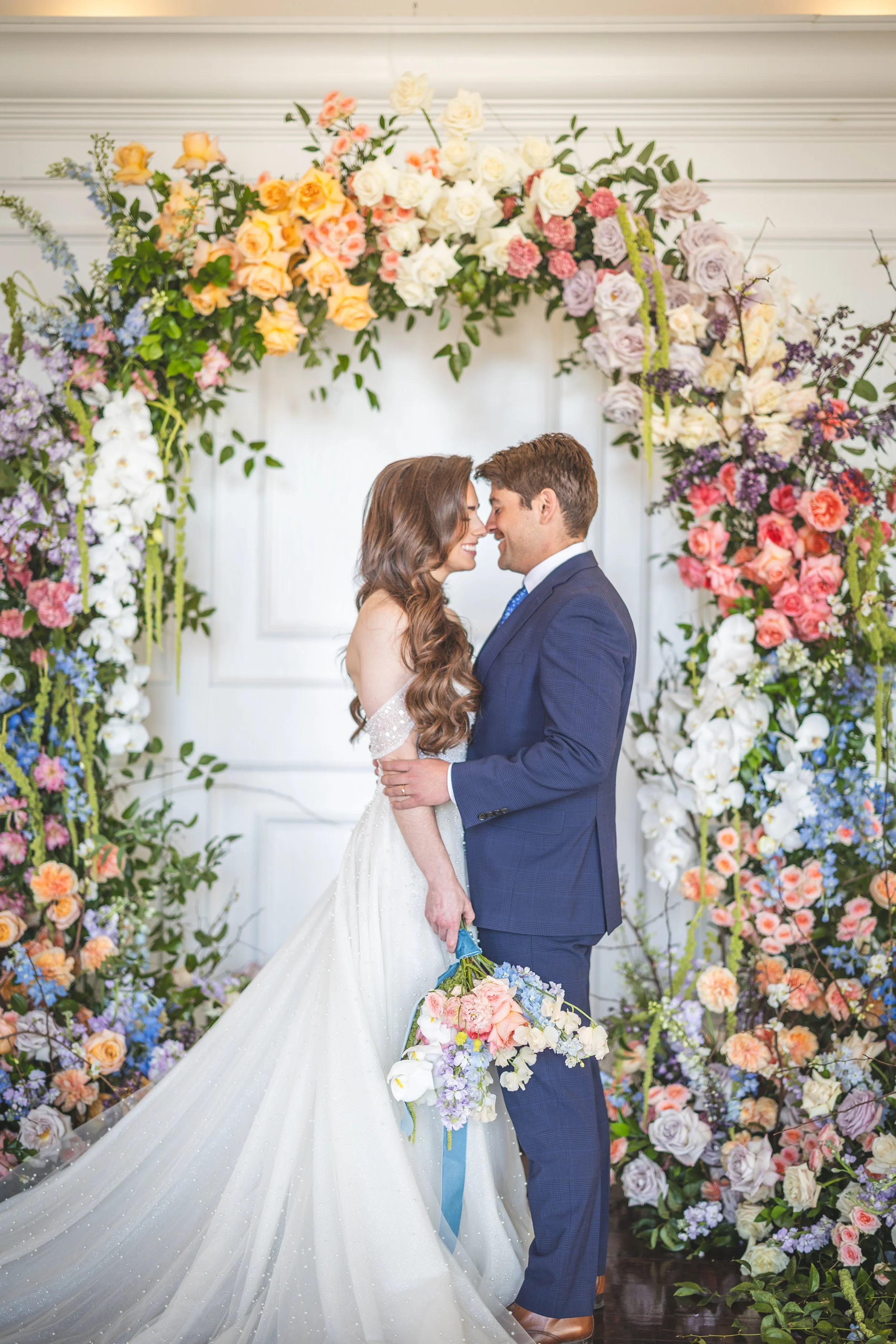 A couple in wedding attire stands close to each other, touching foreheads and smiling at each other, with a large floral arch in the background. The bride wears a white gown with a long, sparkly train, and the groom is dressed in a navy blue suit. Th