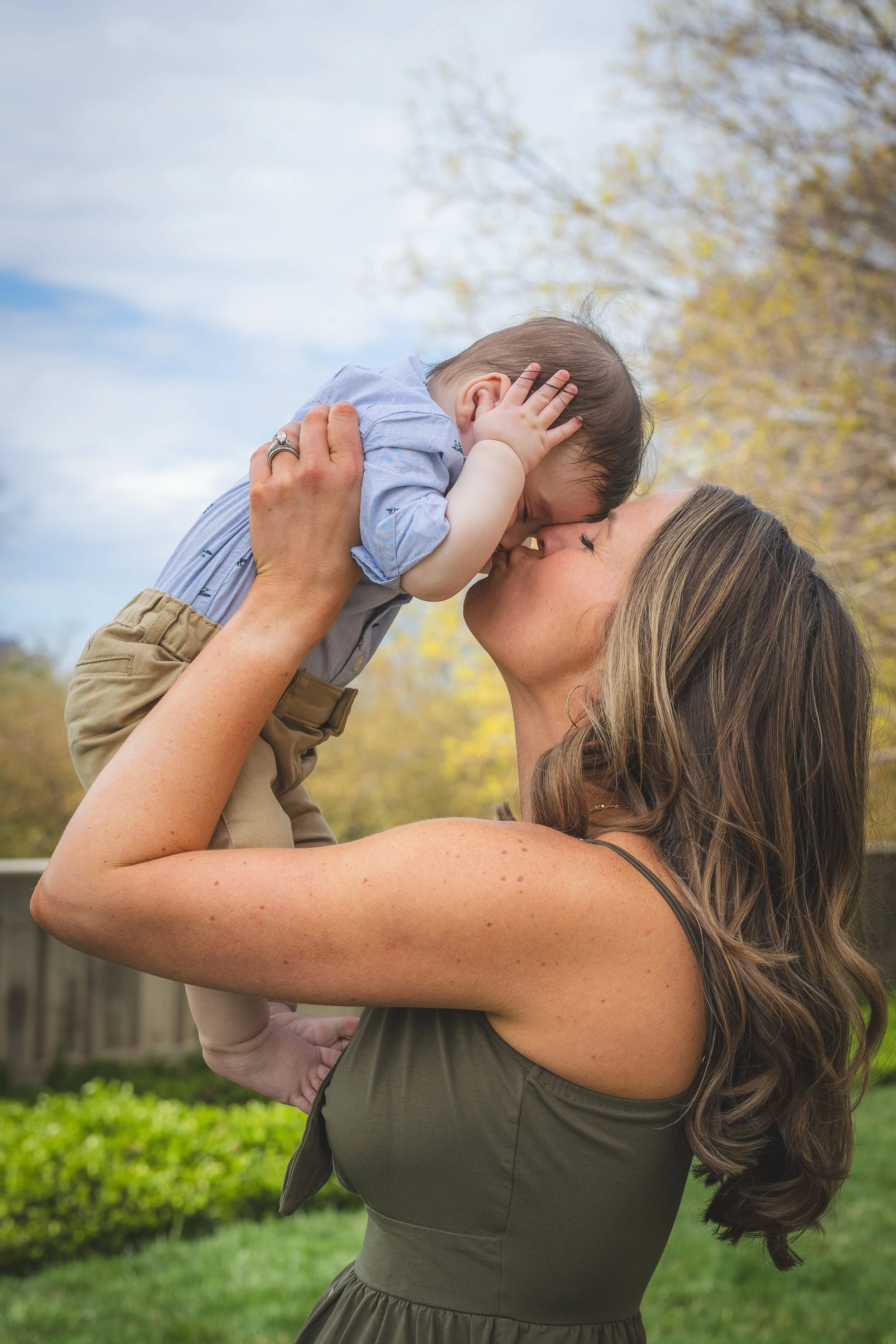 A woman holding a young boy up and giving him a kiss outdoors, with trees and a wooden fence in the background.