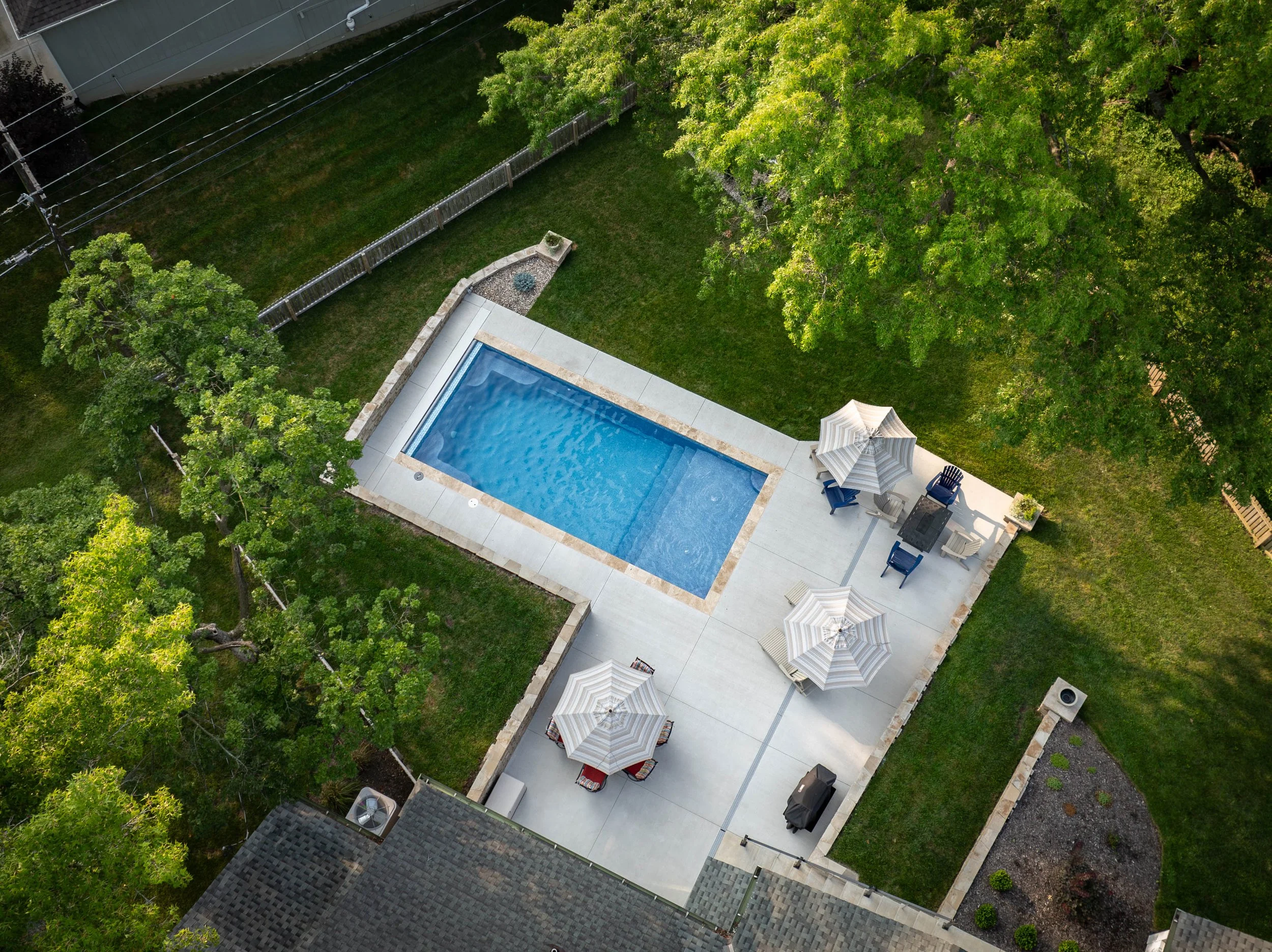 An aerial view of a backyard with a swimming pool, outdoor furniture including umbrellas, chairs, and a fire pit, surrounded by green trees and grass, with a wooden fence enclosing the yard.