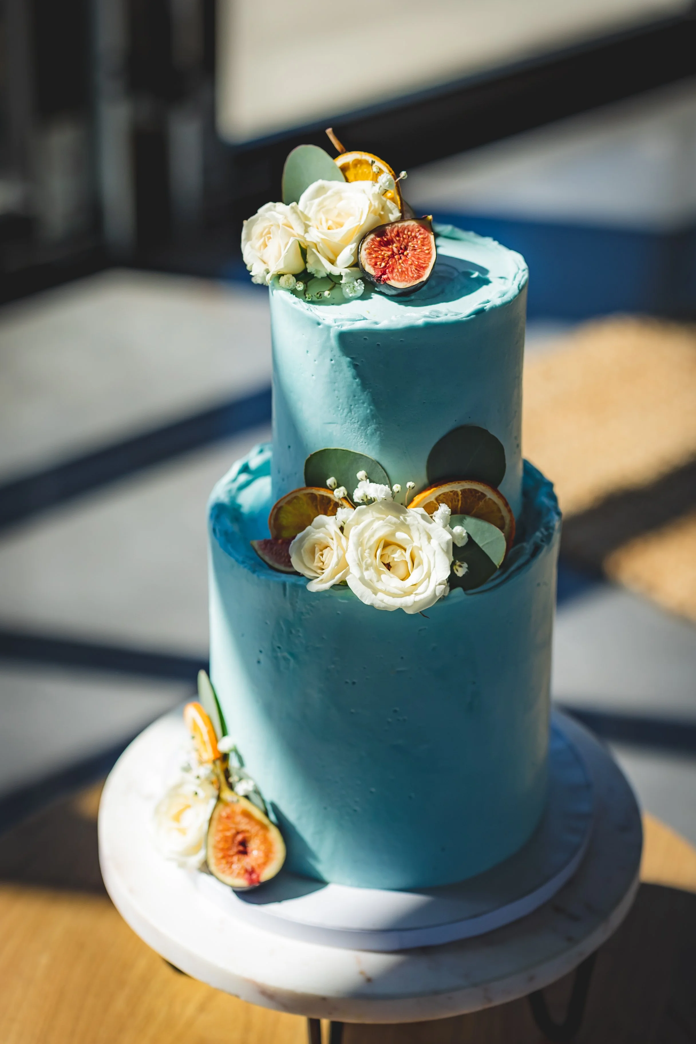 A two-tiered blue wedding cake decorated with white roses, figs, and citrus slices on top and around the base.