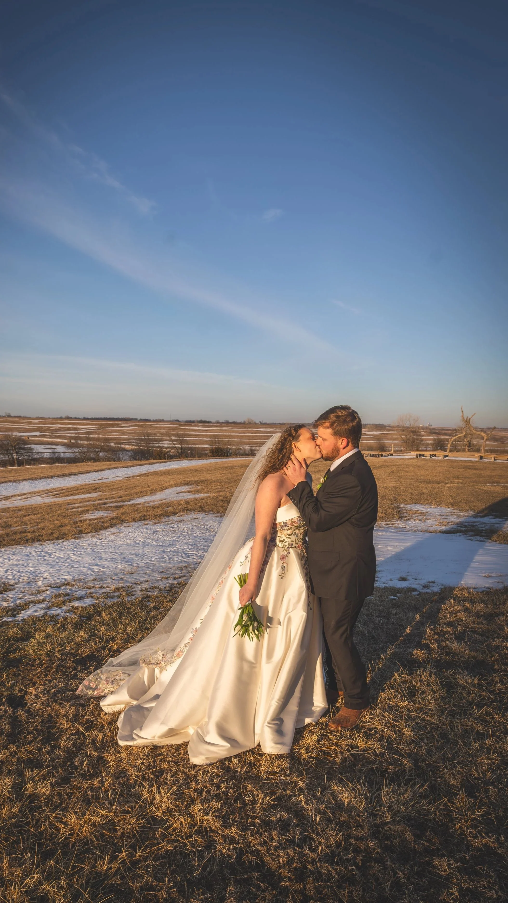 A couple in wedding attire sharing a kiss outdoors on a partly snowy field under a blue sky.