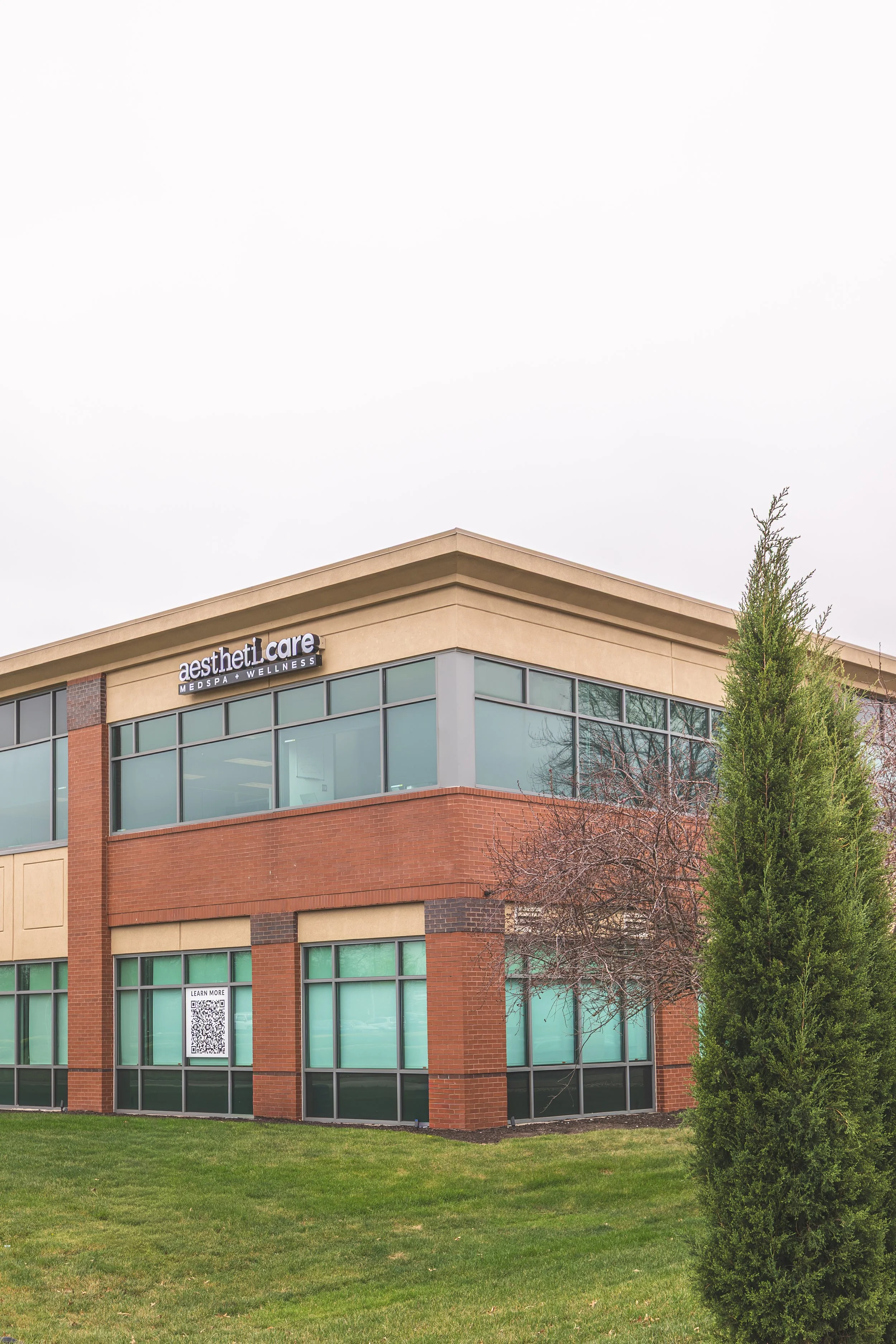 A modern, multi-story commercial building with a beige upper facade and red brick lower section. The building has large glass windows and a sign that reads 'aesthetic care MedSpa Wellness.' There is a green lawn in the foreground, a tall evergreen tr