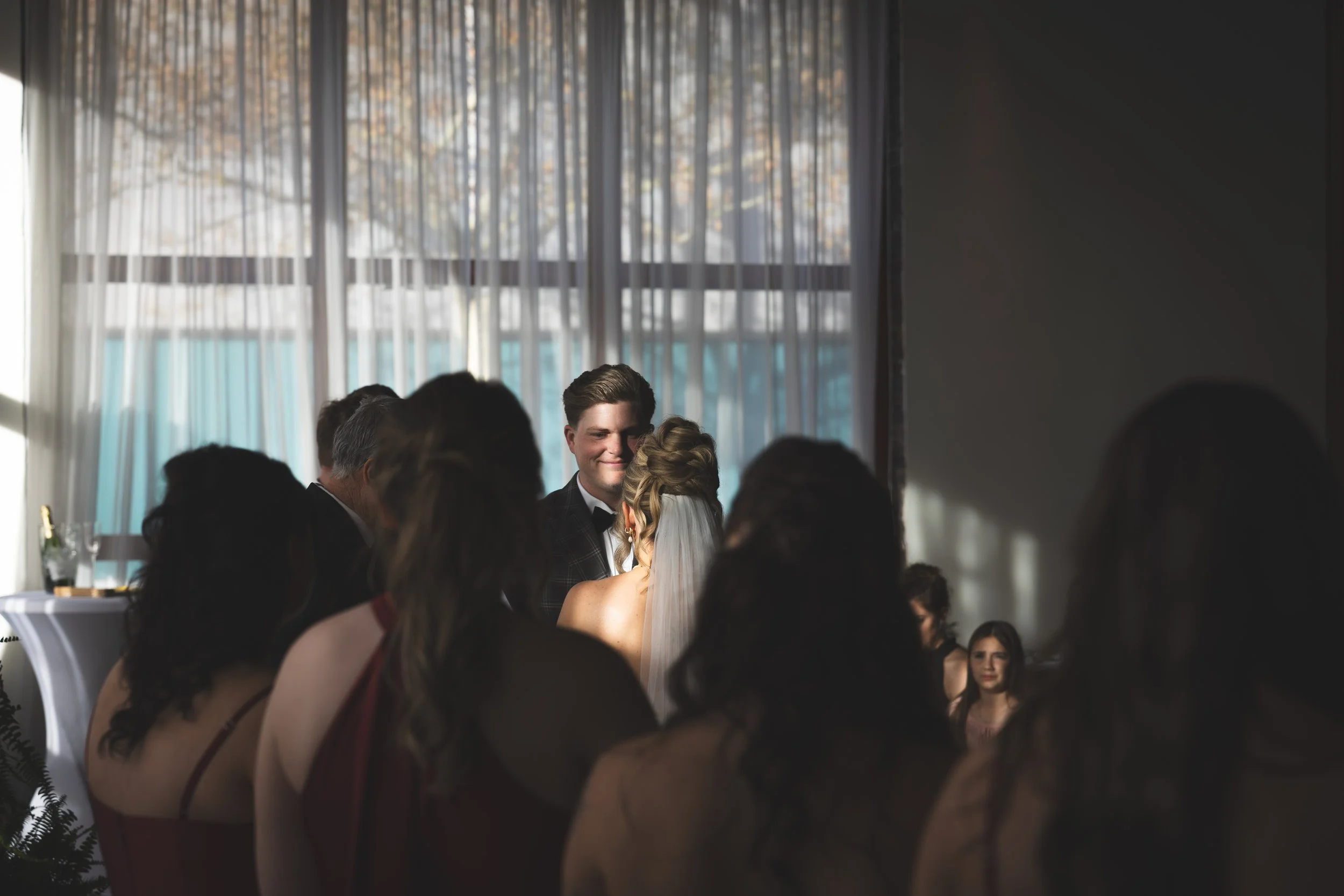 A wedding ceremony taking place indoors with a bride and groom smiling at each other, surrounded by family and friends, with large windows in the background letting in natural light.
