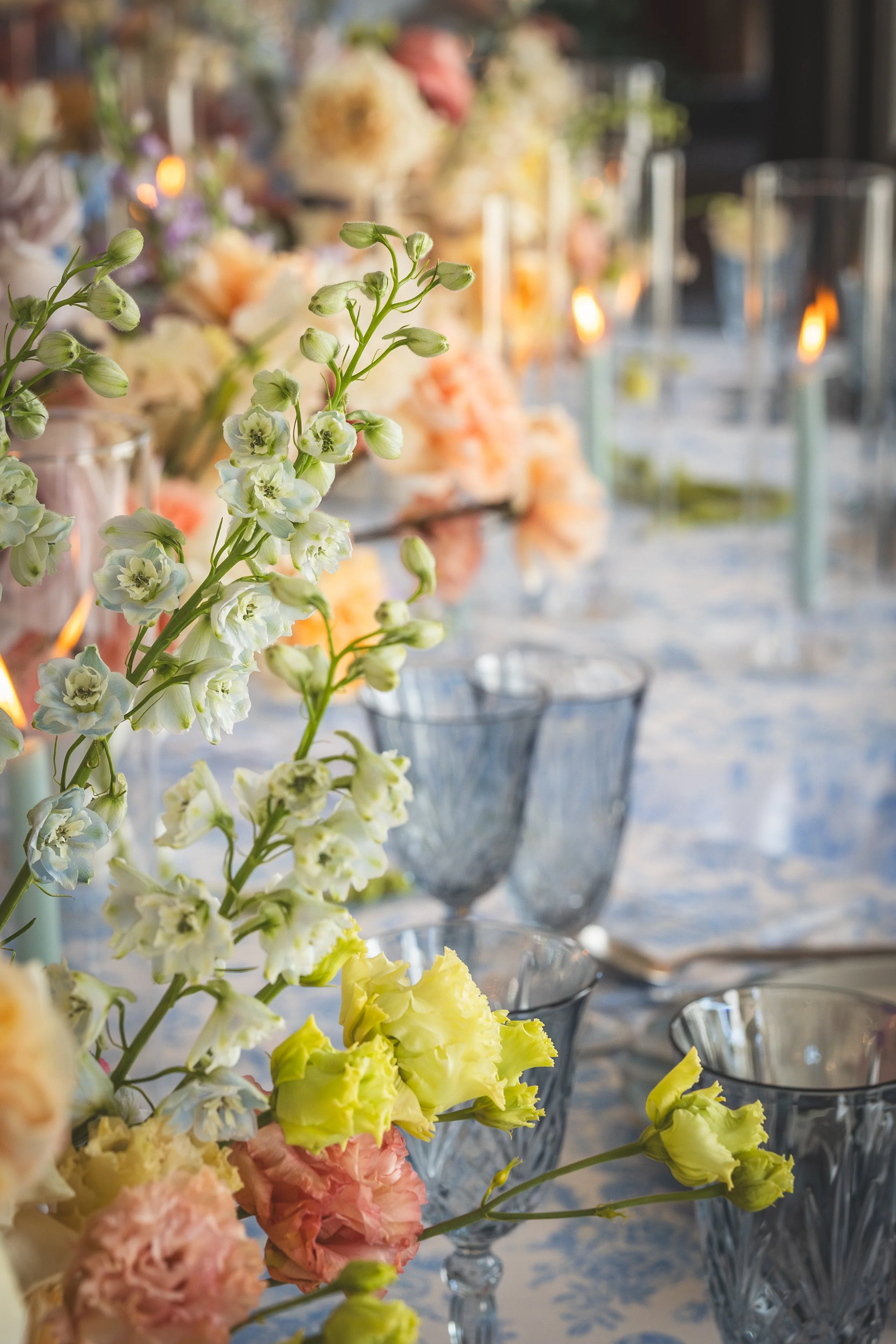 Close-up of a floral table centerpiece with pastel-colored flowers, including white, yellow, light pink, and peach blooms, and empty glass stemware positioned on a table with a floral-patterned tablecloth. Smoldering candles in glass holders are in t