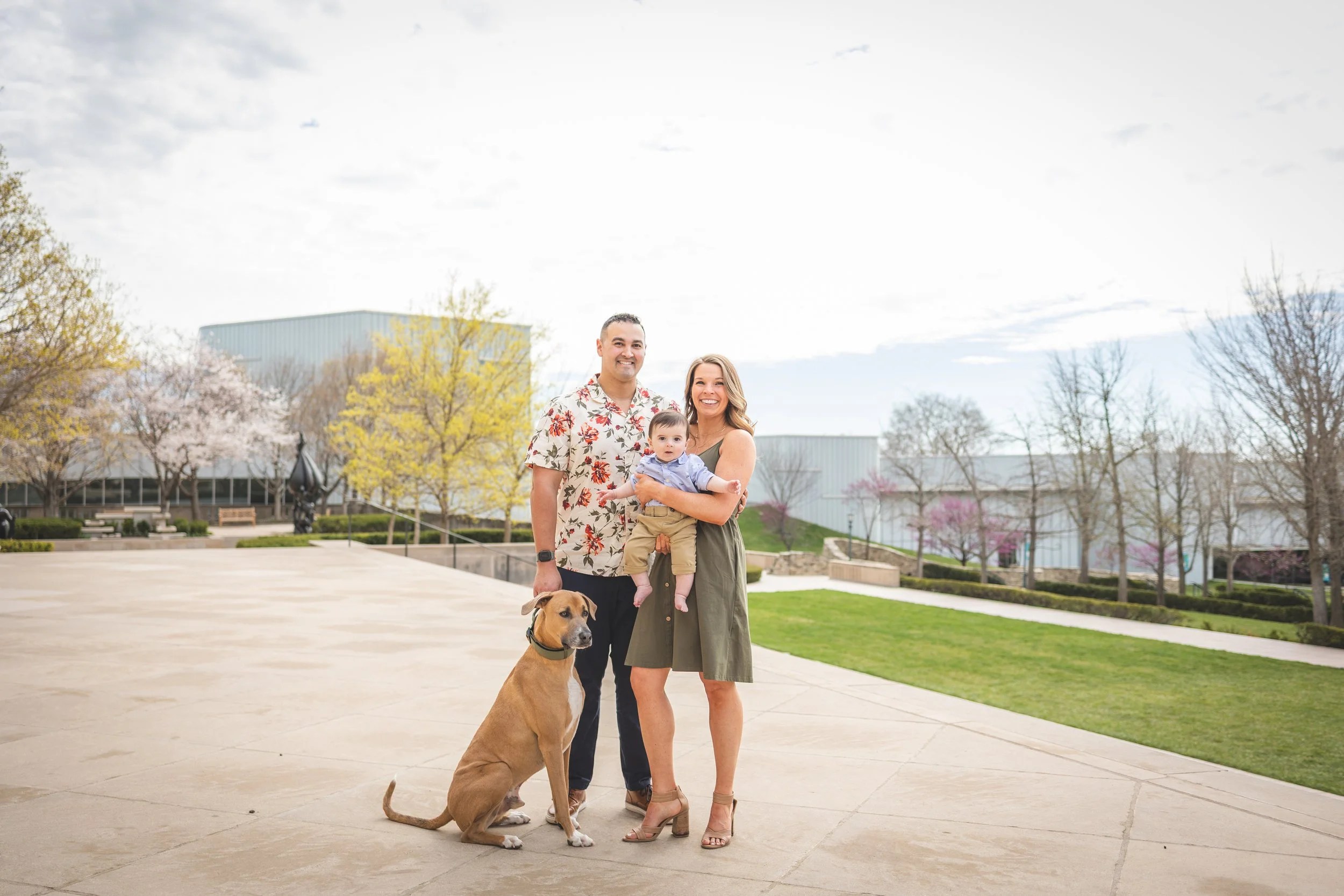 A family of three with a dog posing outdoors in a park with spring trees and buildings in the background.