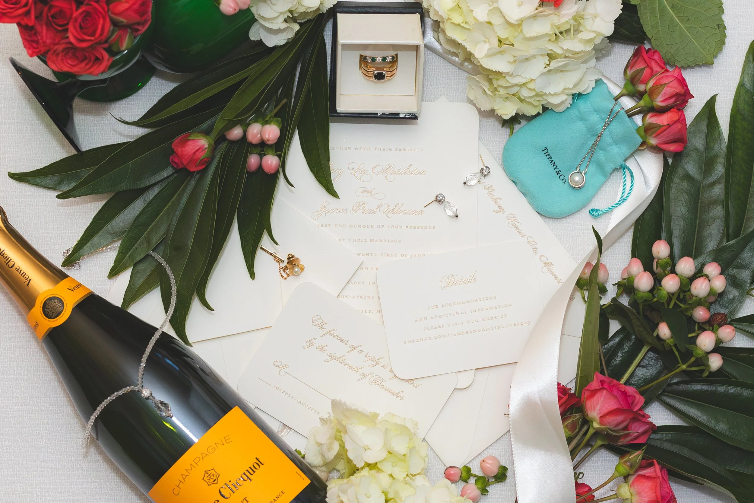 A wedding invitation suite surrounded by flowers, jewelry, a champagne bottle, and Tiffany & Co. box, all arranged on a white table