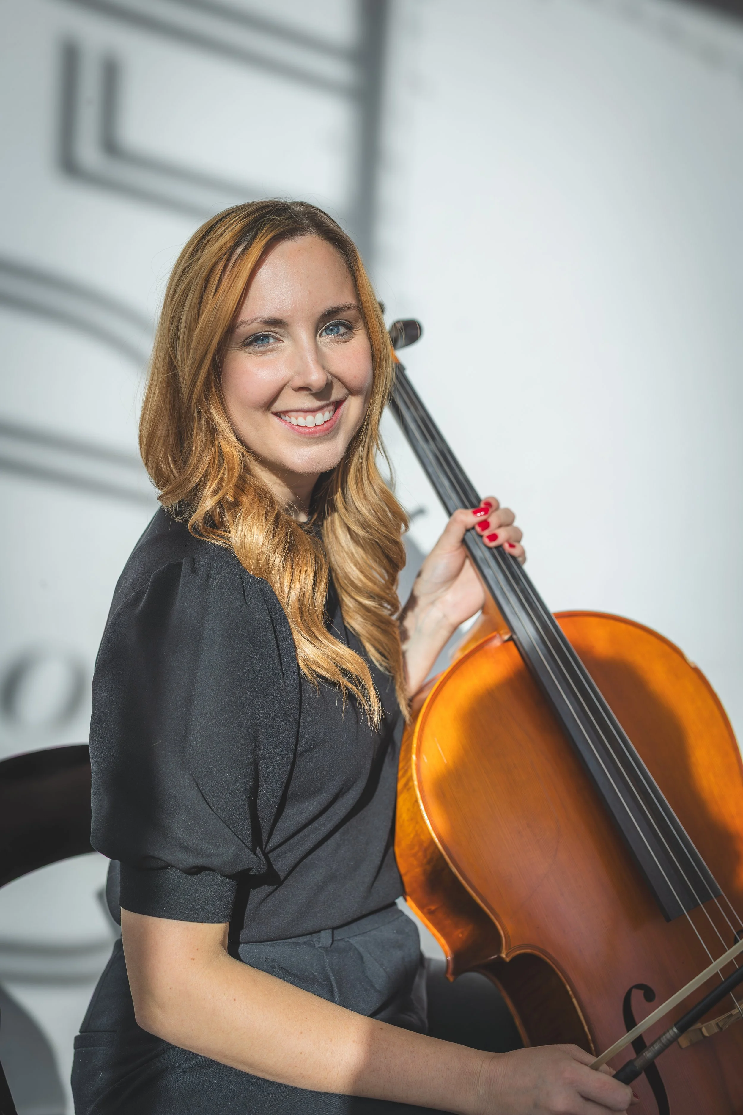 A woman with red hair and blue eyes smiling while holding a cello and a bow, wearing a black dress, in a room with blurred background.