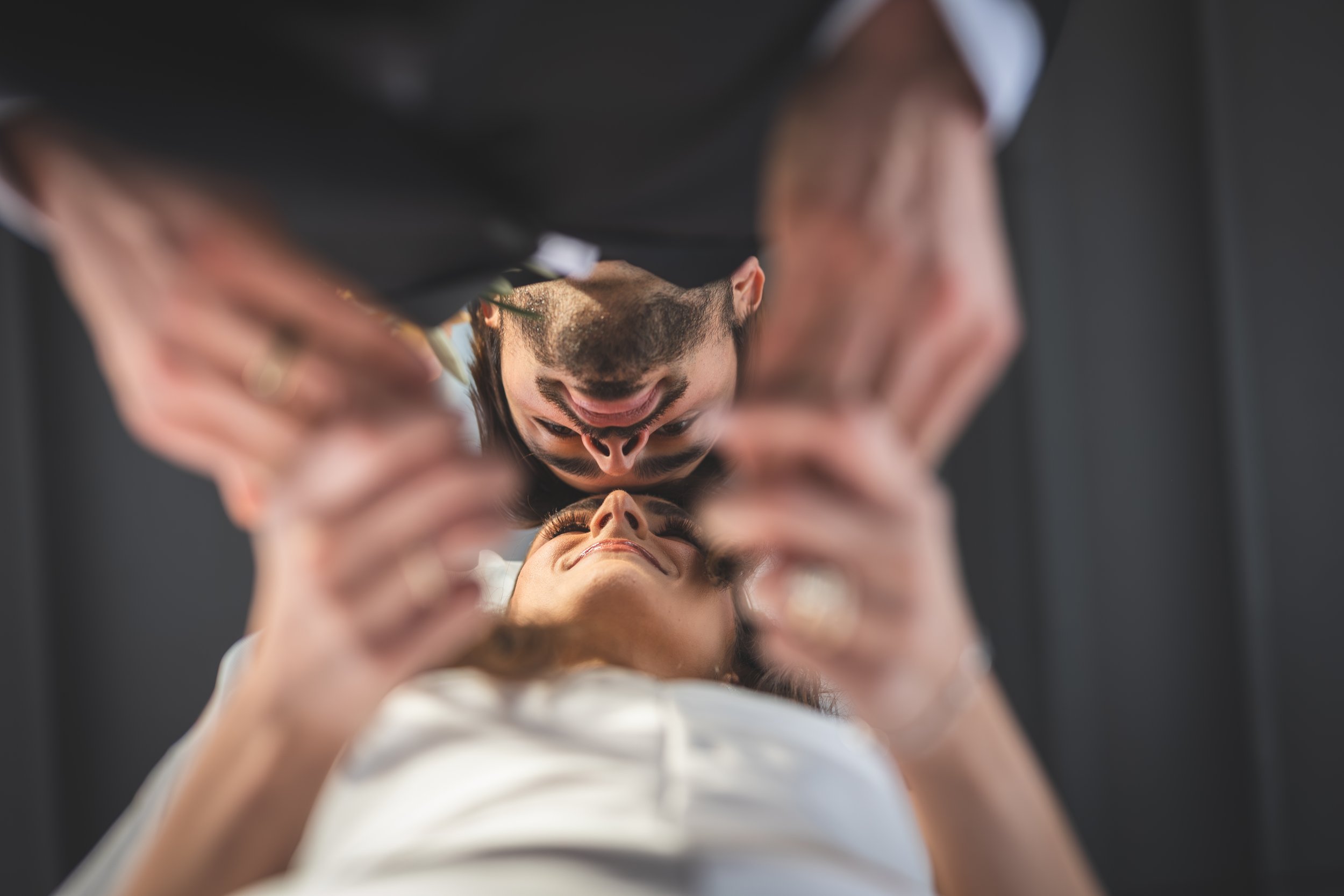 A person with a beard and mustache is looking at another person's face while holding a mirror in front of them, captured from below with a dark background.