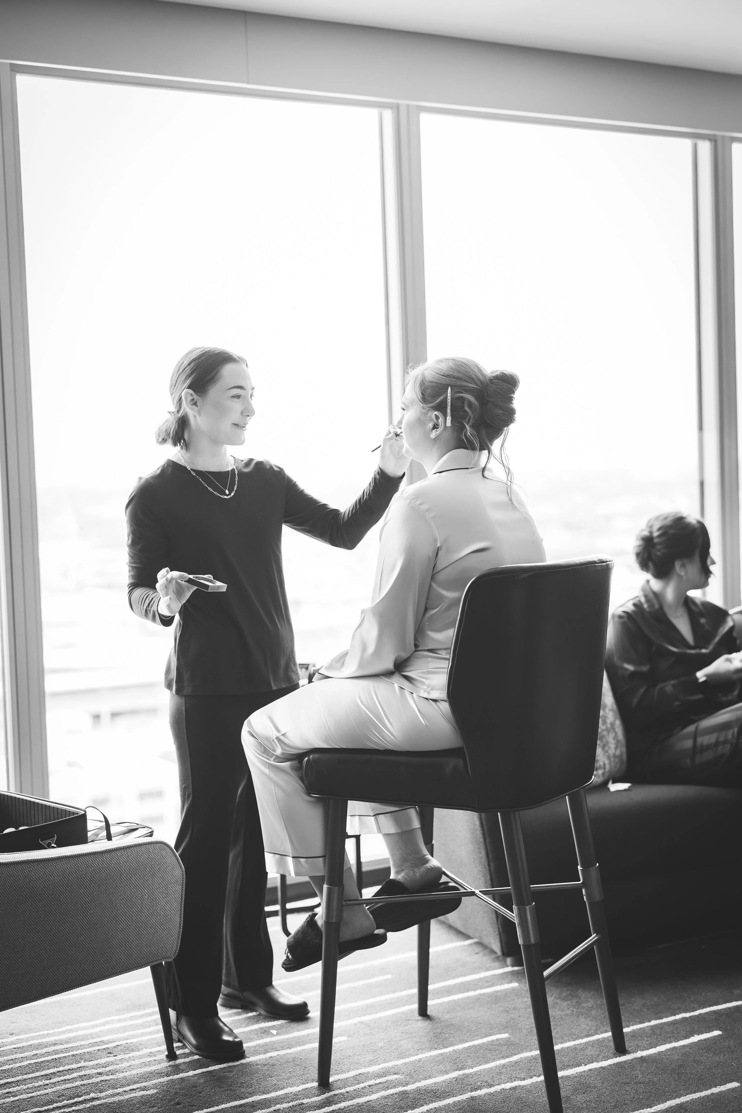 A woman sitting on a high chair by a window, getting her makeup done by a makeup artist standing in front of her. Two women are visible in the background, one furthest from the window looking down and the other in the corner.