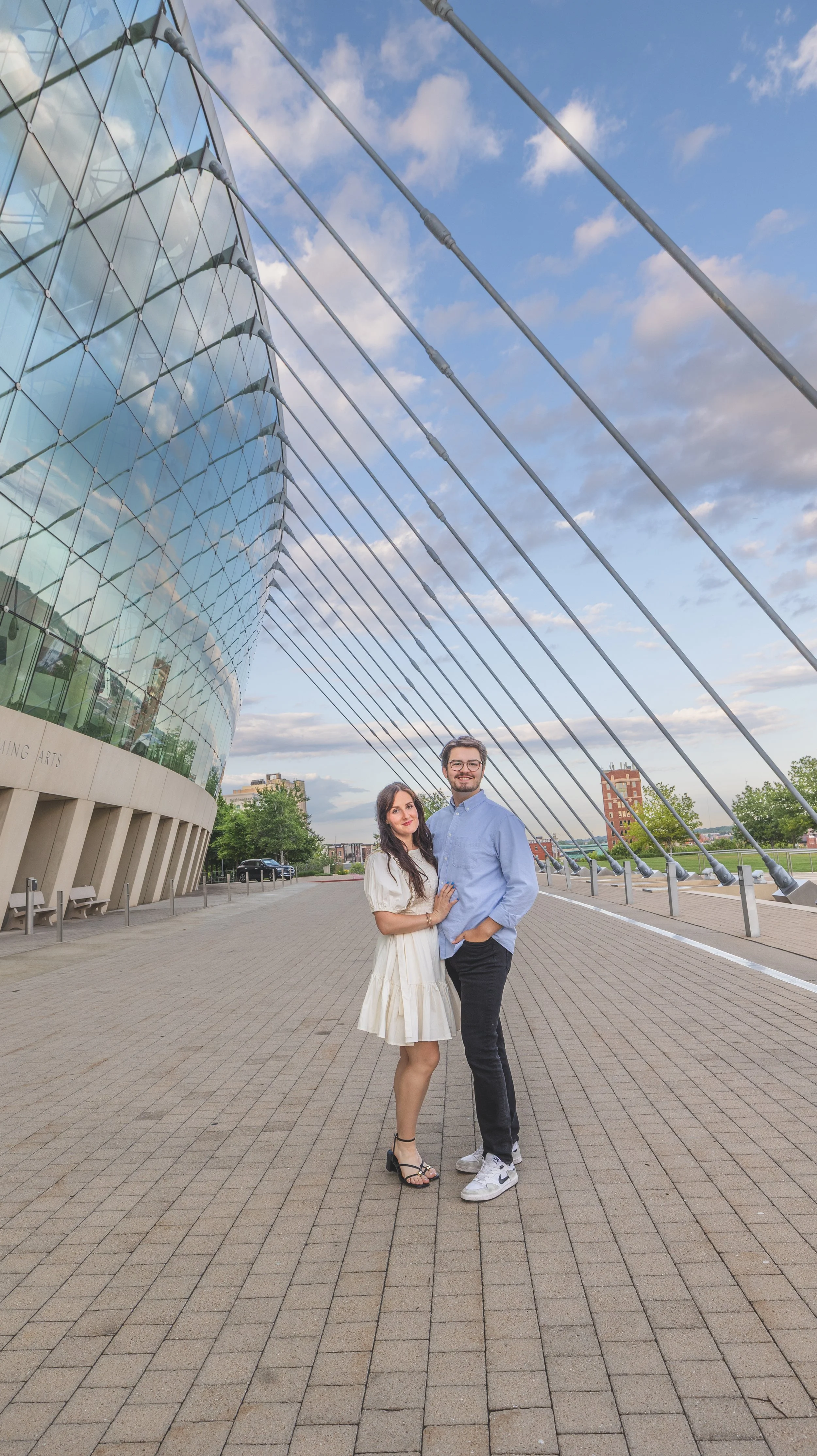 A young couple standing close together outside in front of a modern glass building with metal support beams and a cloudy sky, smiling at the camera.