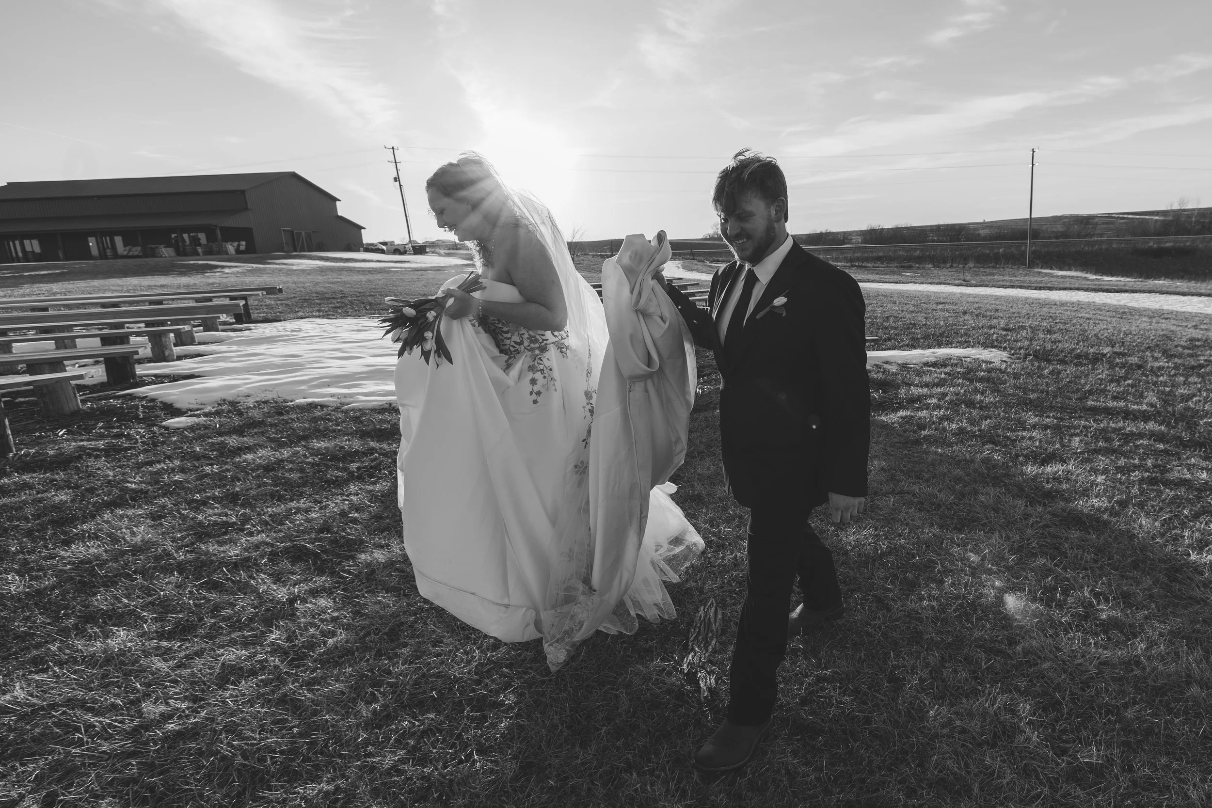 A black and white photo of a wedding scene outdoors, with a bride holding her wedding gown and a bouquet, and a groom walking beside her, both smiling under the sunlight with a building and power lines in the background.
