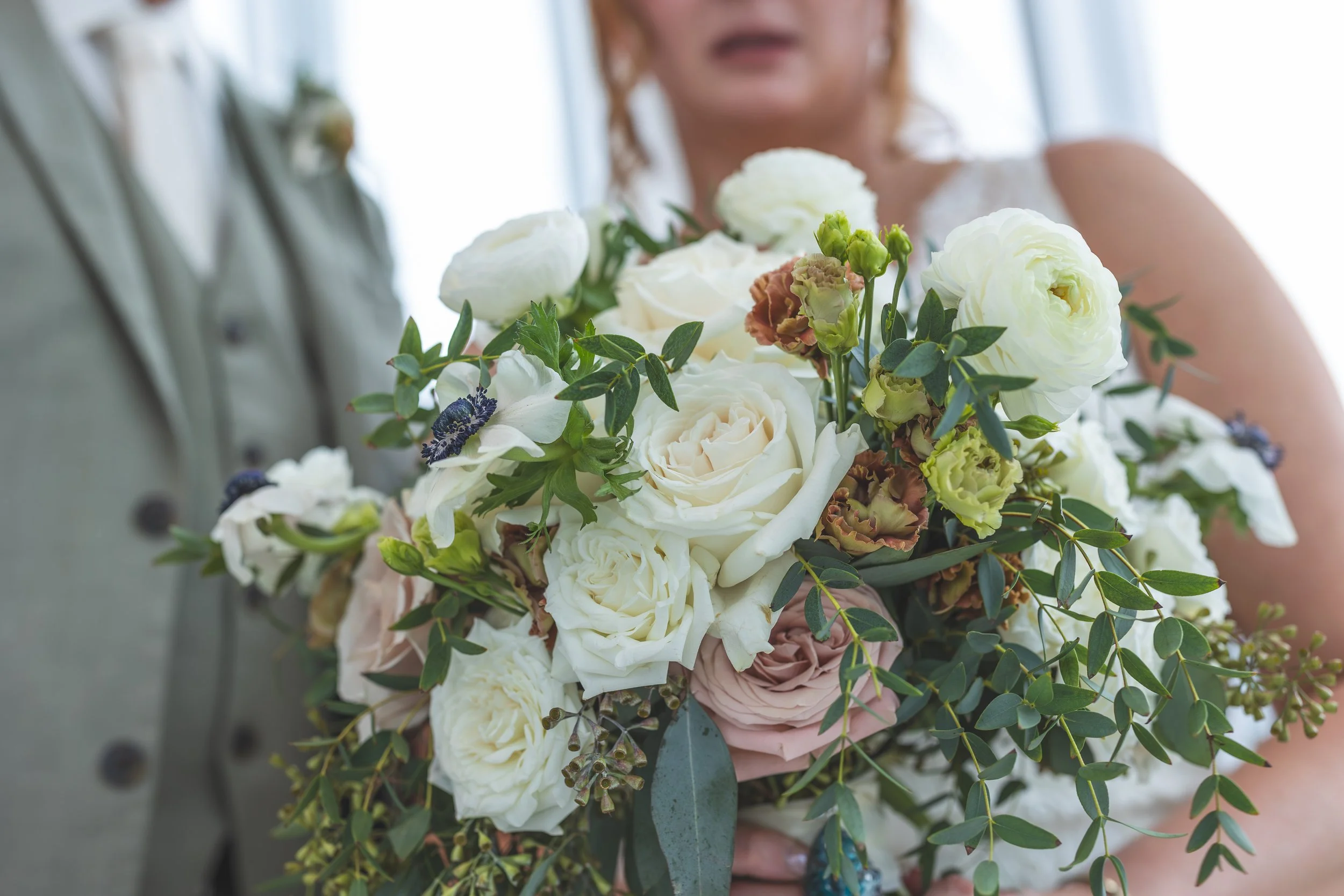 Close-up of a woman holding a bouquet of white and pink roses with green foliage, with a blurry man in the background.
