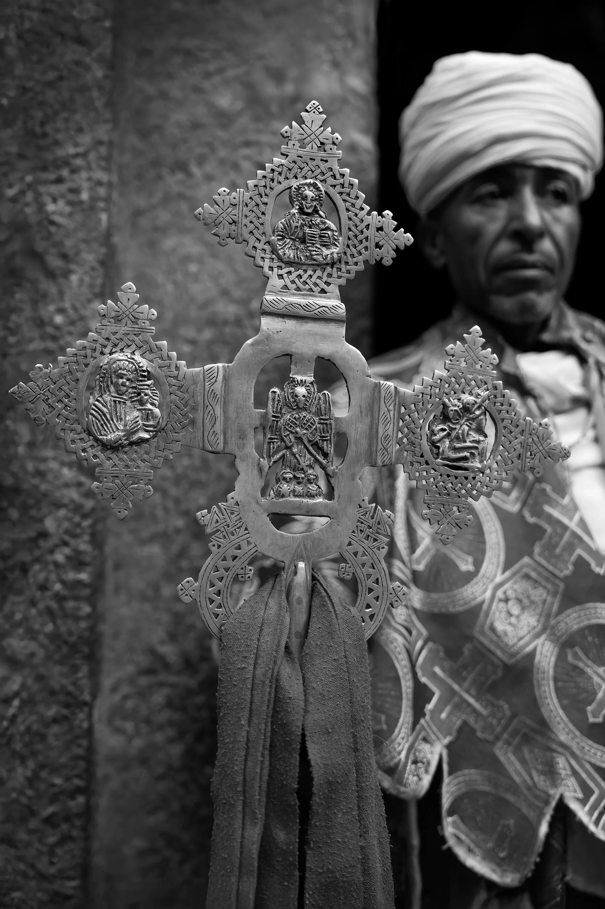 A detailed metallic cross held by a person dressed in traditional clothing, with a blurred background of a stone wall and a woman wearing a headwrap.