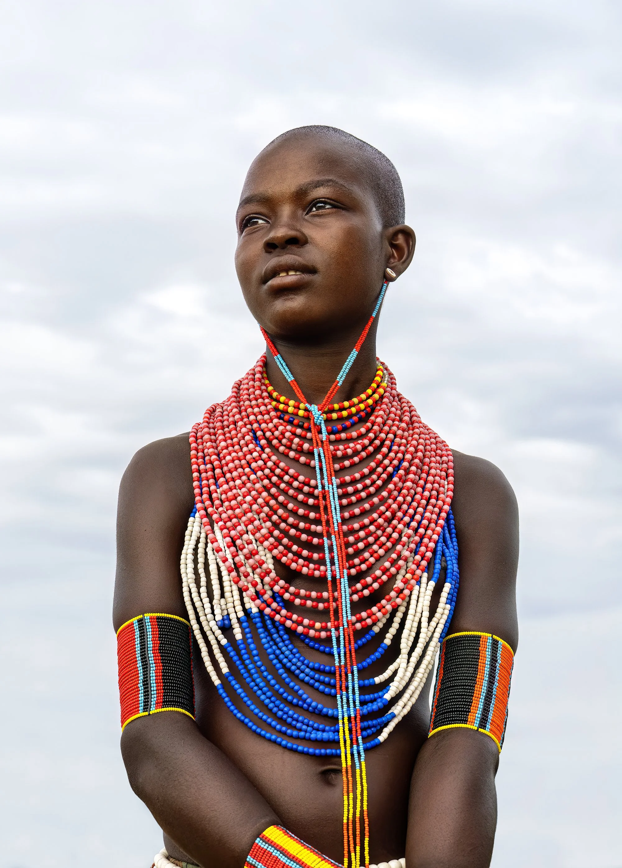Young person with dark skin wearing traditional Maasai jewelry, including multiple layered colorful beaded necklaces, armbands, and earrings, standing outdoors against a cloudy sky.
