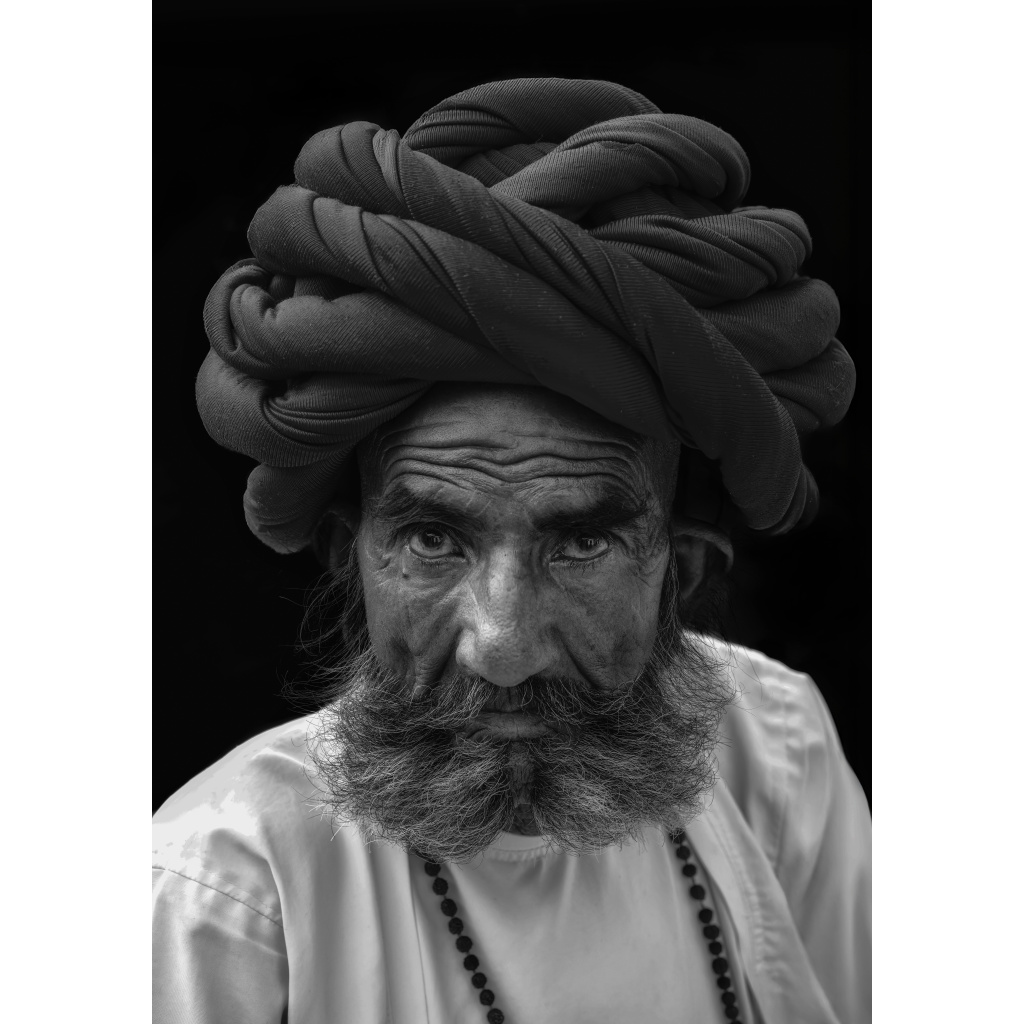 Black and white portrait of an elderly Rabari man wearing a large coilded turban and beaded necklace. His steady gaze and textured skin reveal a lifetime shaped by the desert light of Rajasthan.