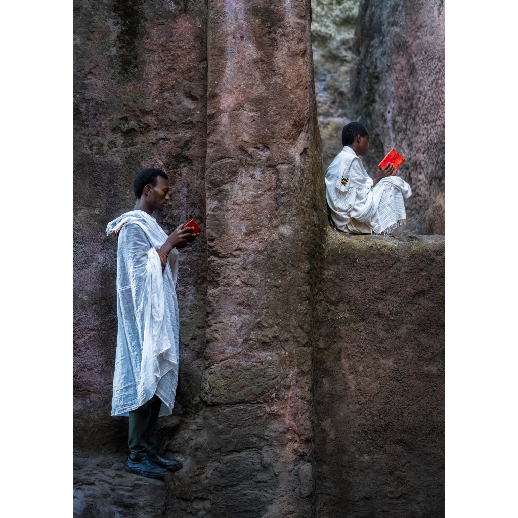 Two young men wearing white traditional clothing, sitting and standing on rocky cliffs while reading red books.