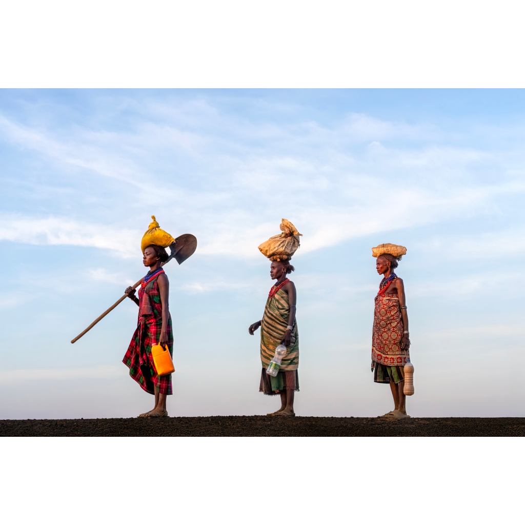 Three women in traditional attire walking in a line on a dirt path under a blue sky, carrying goods on their heads and bottles in hand.