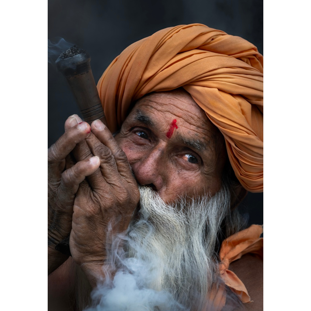 A Rabiri elder in Rajasthan holds a chillum close to his lips.  His orange turban and deep, lined expression are softly lit against a dark background, capturing a moment of still reflection.