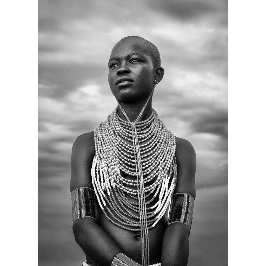A young woman from an African tribe with multiple beaded necklaces, arm bracelets, and a shaved head, looking contemplatively against a cloudy sky.